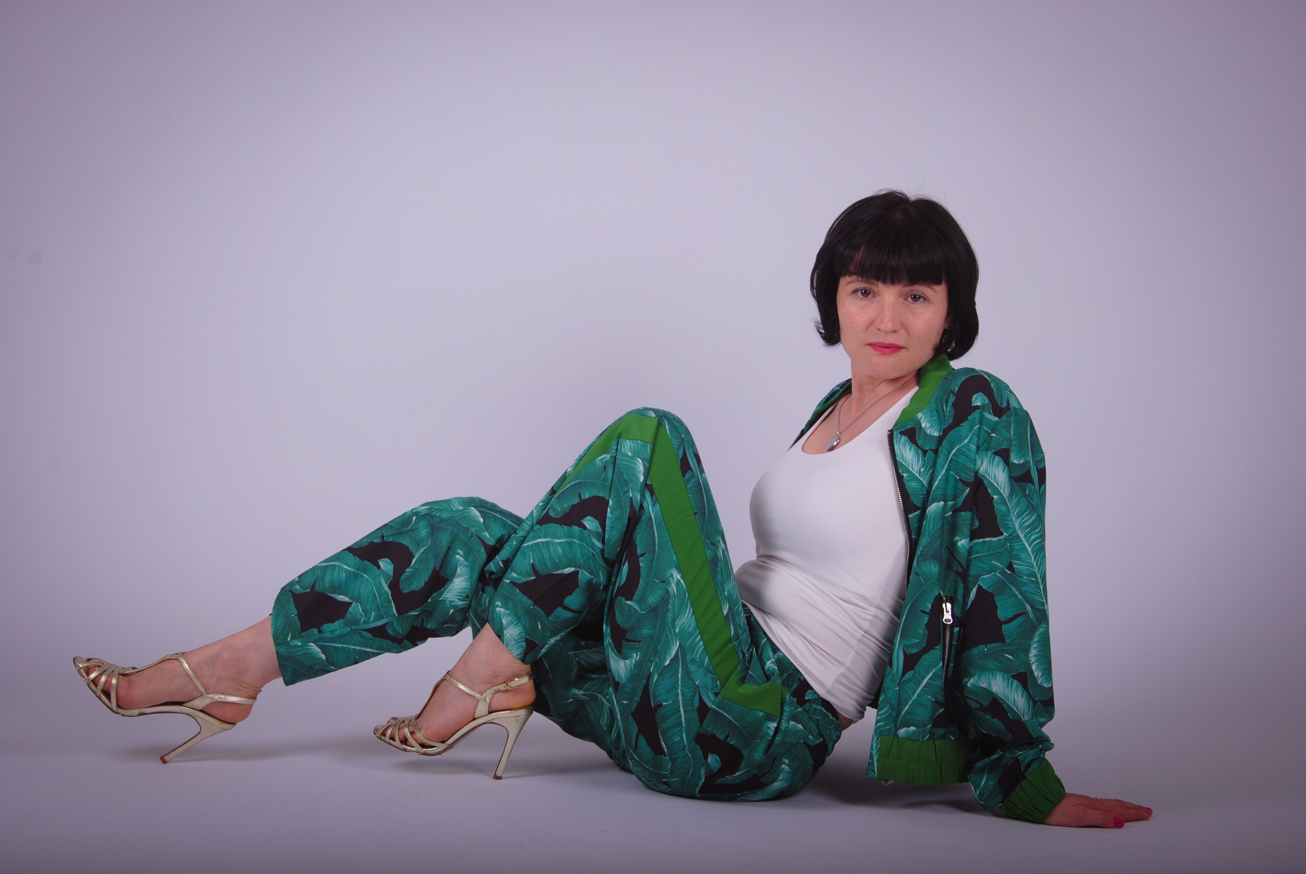 Модель в изумрудном брючном костюме сидит на полу в золотых босоножках 