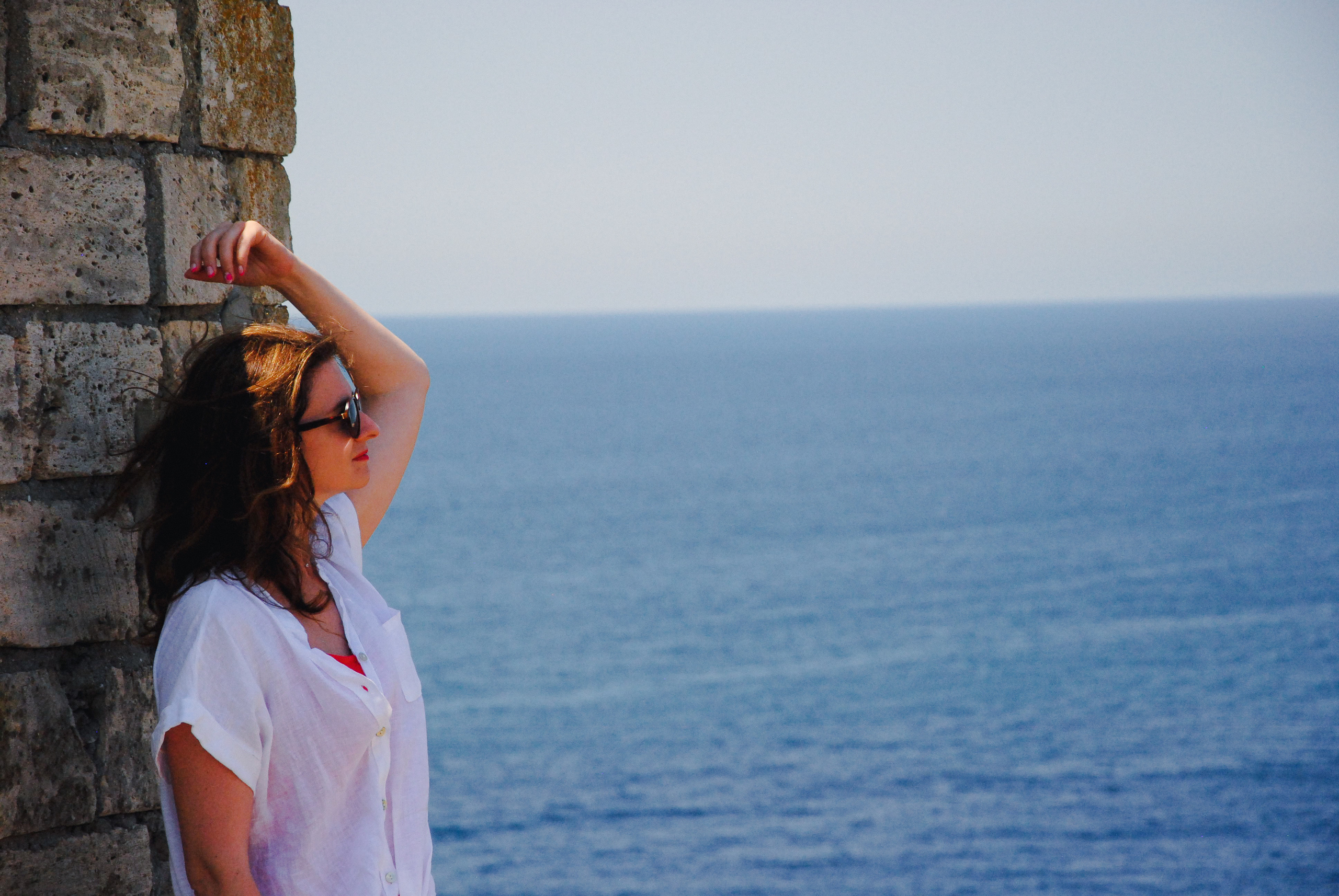 Модель в белой хлопковой рубашке и черных очках стоит возле кирпичной стены на фоне синего моря