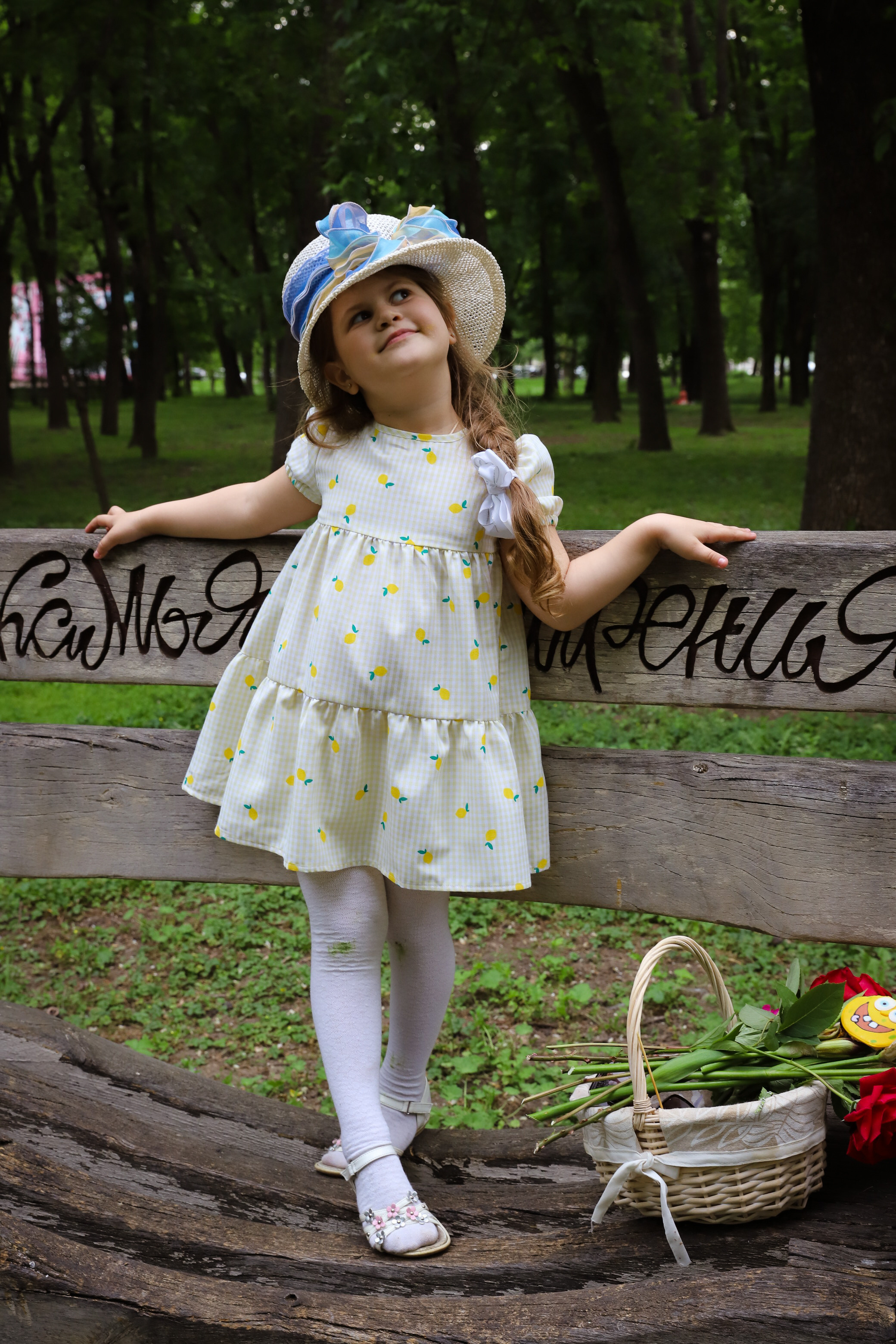 Веселая девочка- хохотушка в шляпе с корзинкой позирует в желтом платье в парке на скамейке