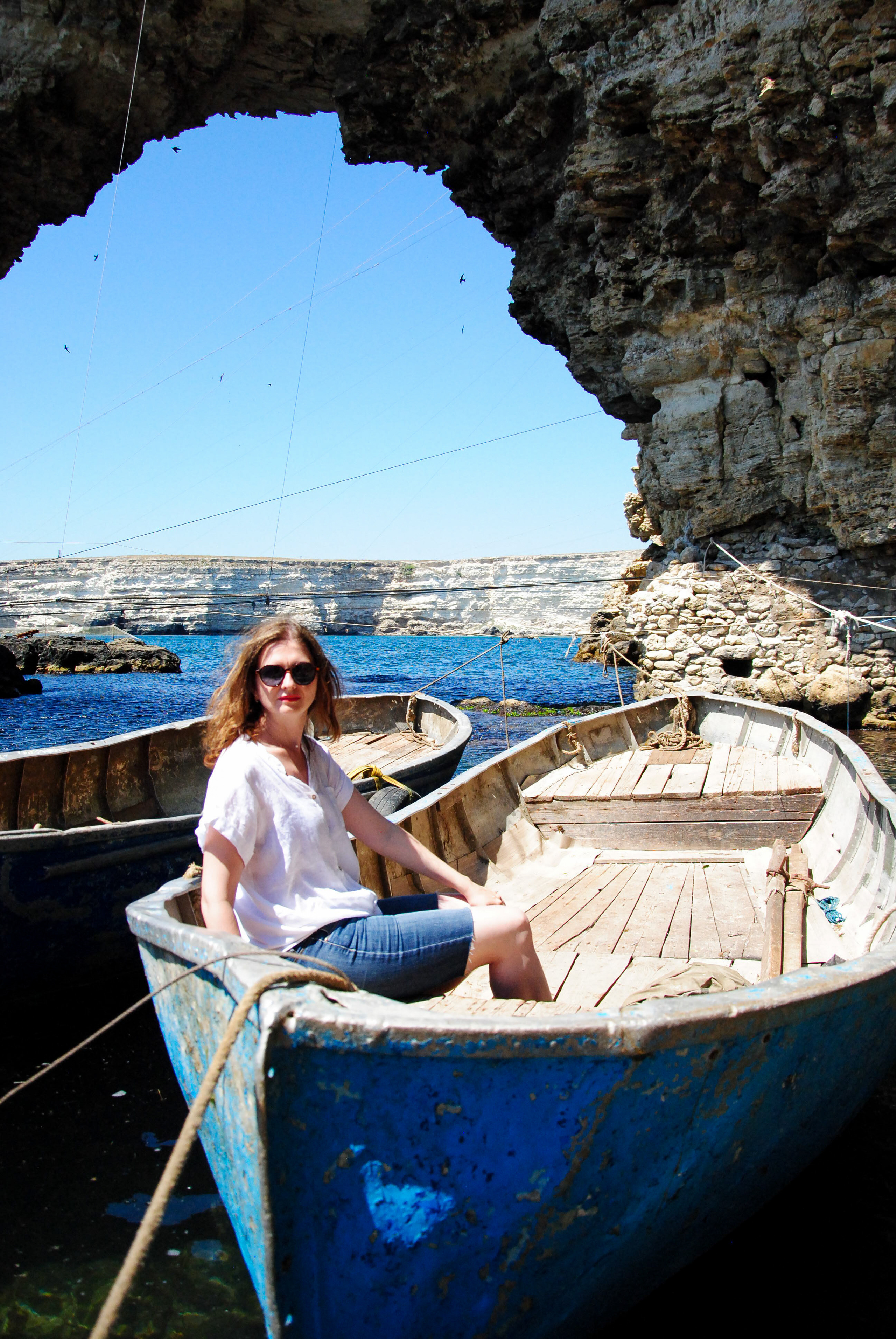 Девушка в белой льняной футболке и джинсовых шортах сидит в деревянной лодке на фоне сквозного  грота в Рыбацком поселке. Малый Атлеш. Крым