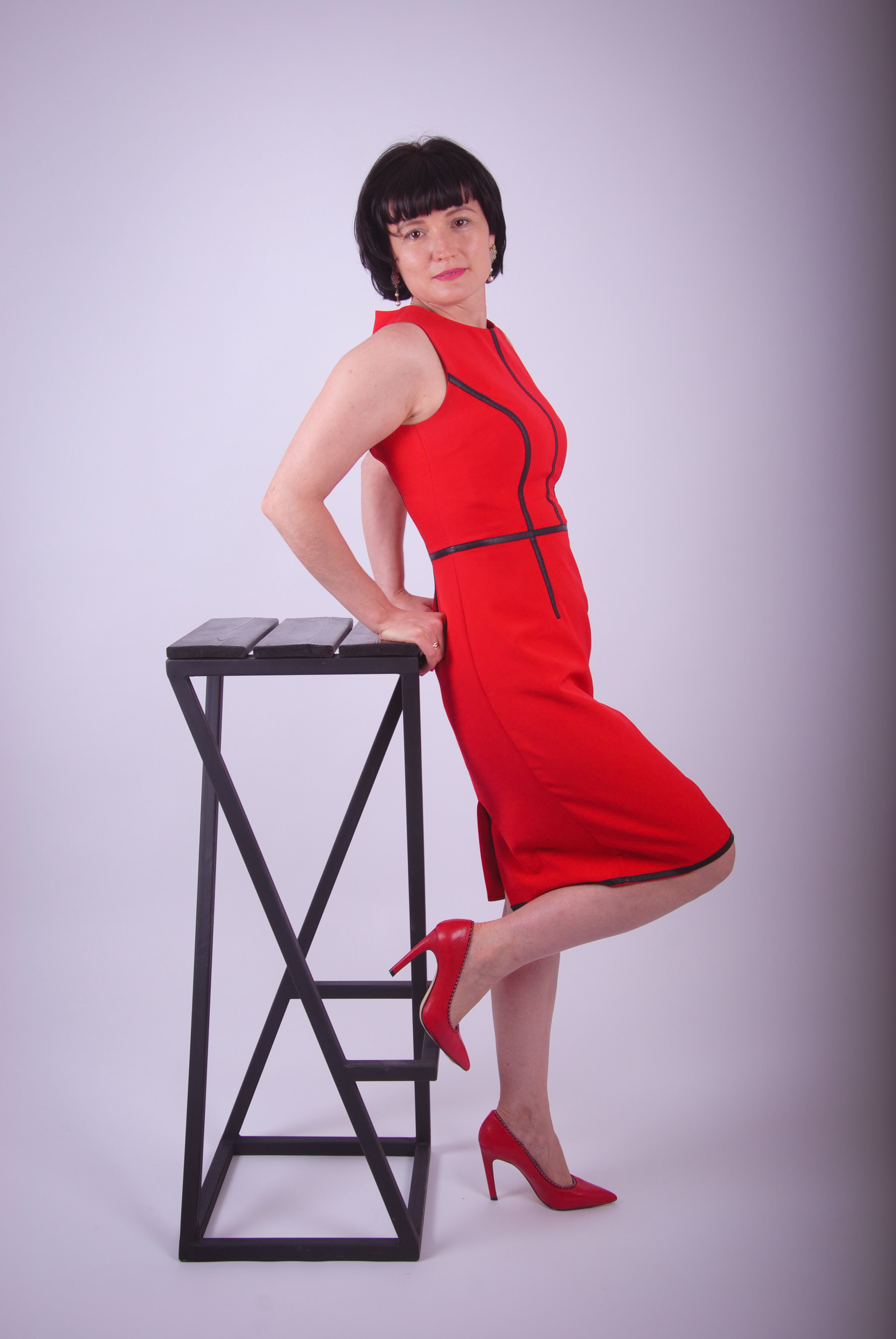 Девушка с темными короткими волосами облокотилась на высокий барный стул в красном платье и в алых туфлях на каблуках