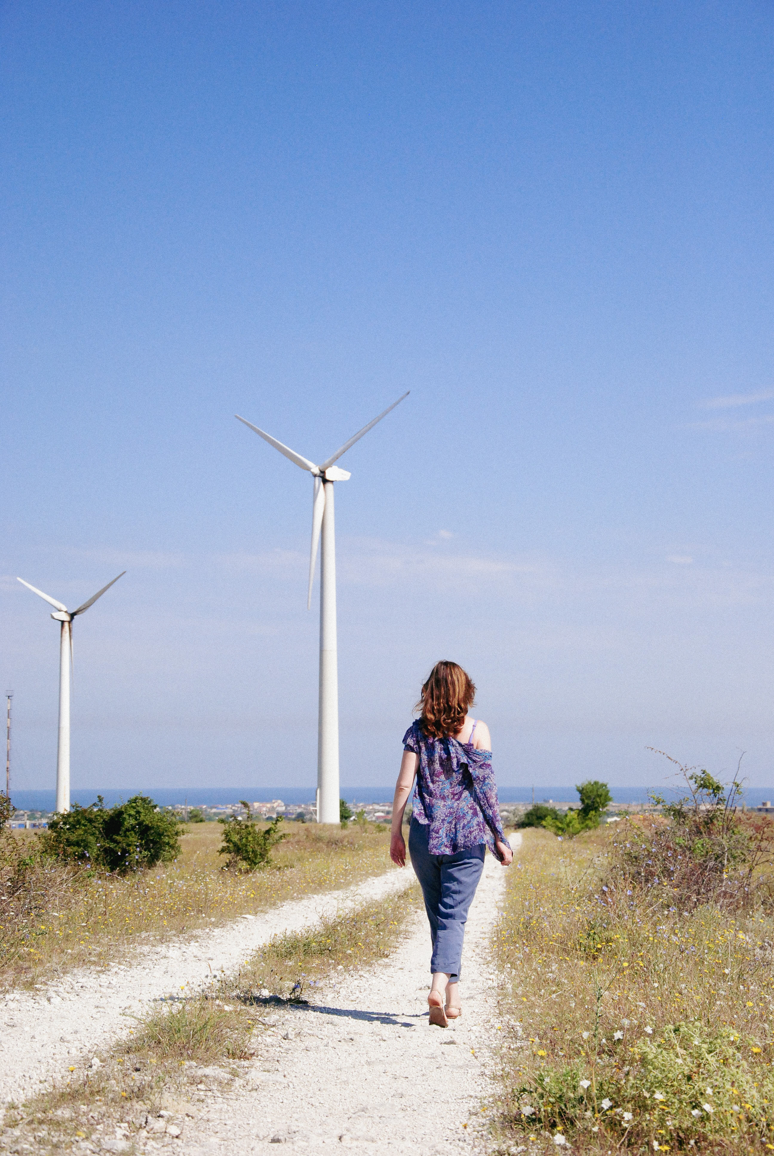 Девушка , уходящая в даль, идет по песчаной дороге на фоне голубого неба и ветряков