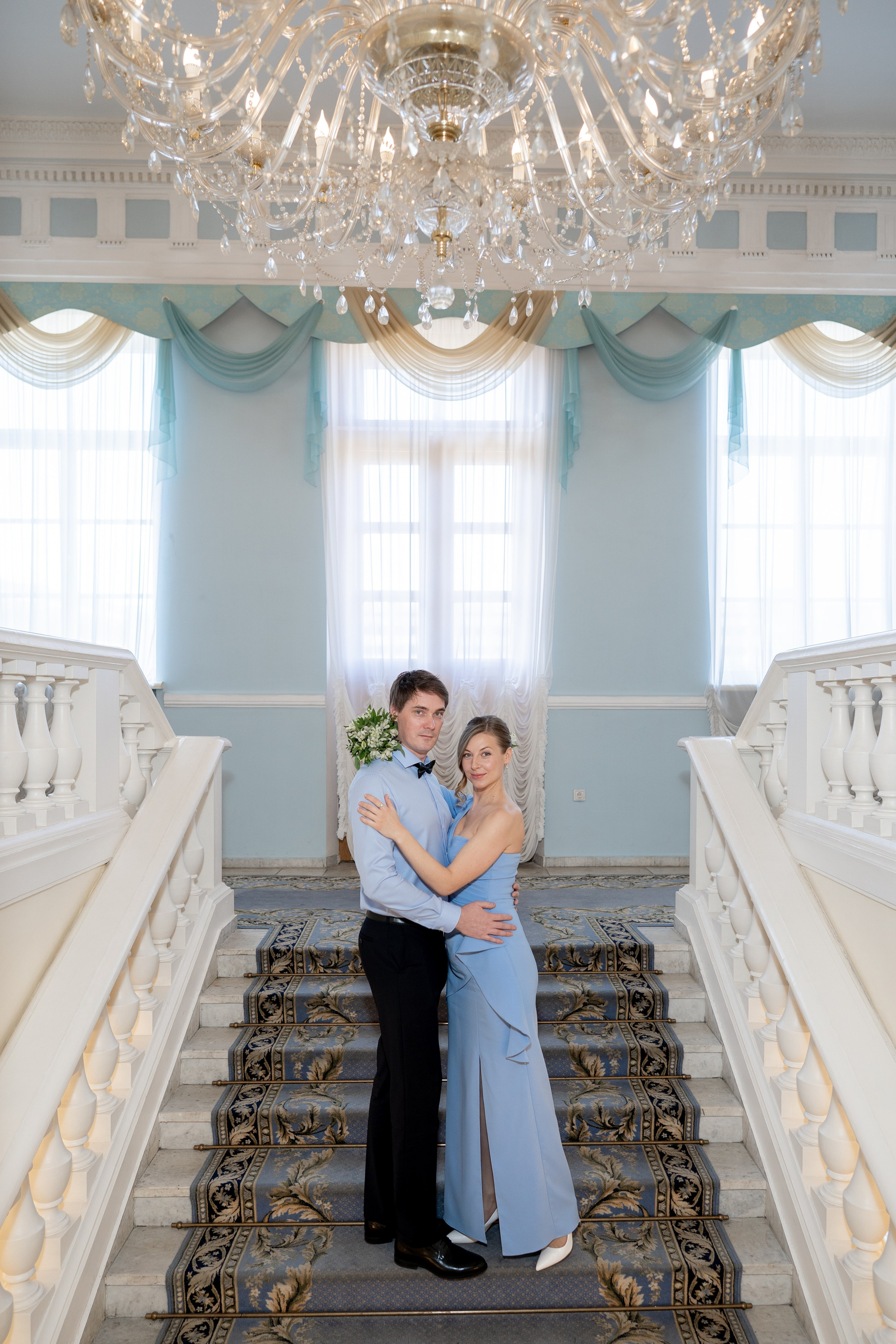 Свадебная фотосессия в ЗАГСе Кировского района. Жених и невеста на парадной лестнице под люстрой обнимаются