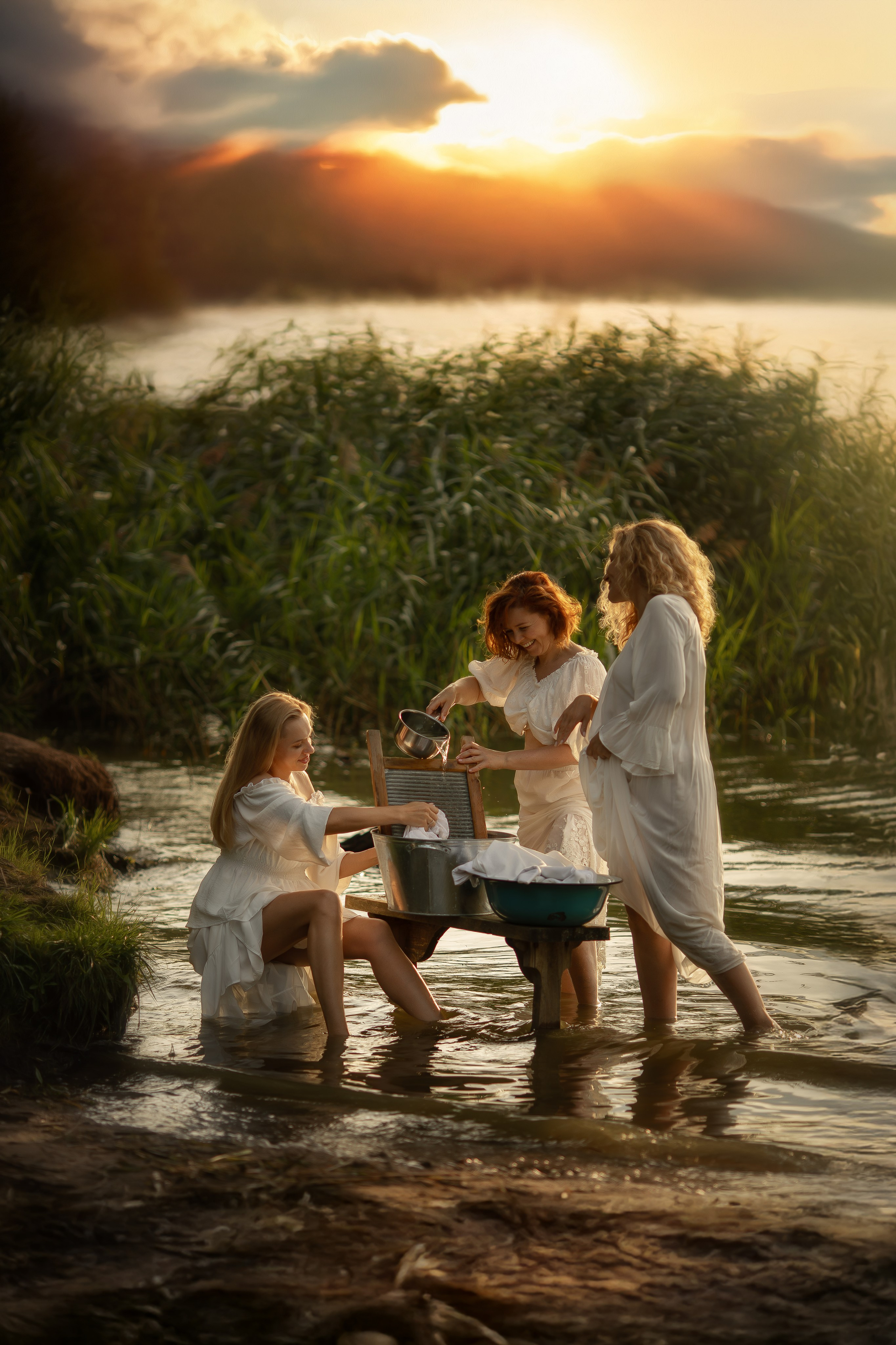 Три девушки в белых платьях стоят в воде у берега озера на закате, стирая бельё в металлическом тазу, вокруг зелёные камыши, над водой мягкий туман и золотые лучи солнца.