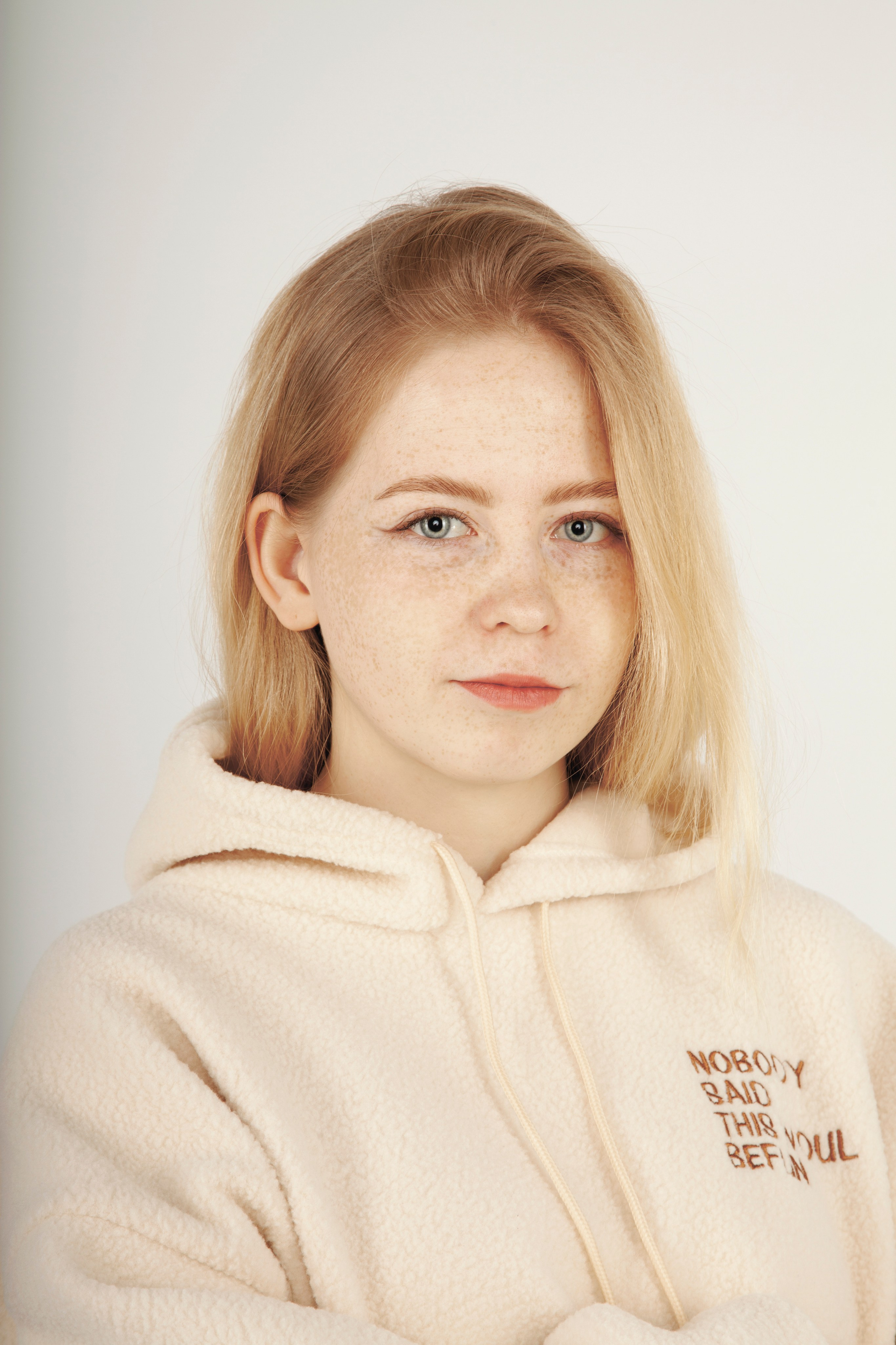 Портрет светловолосой и голубоглазой девушки с веснушками. В светлой одежде на белом фоне.