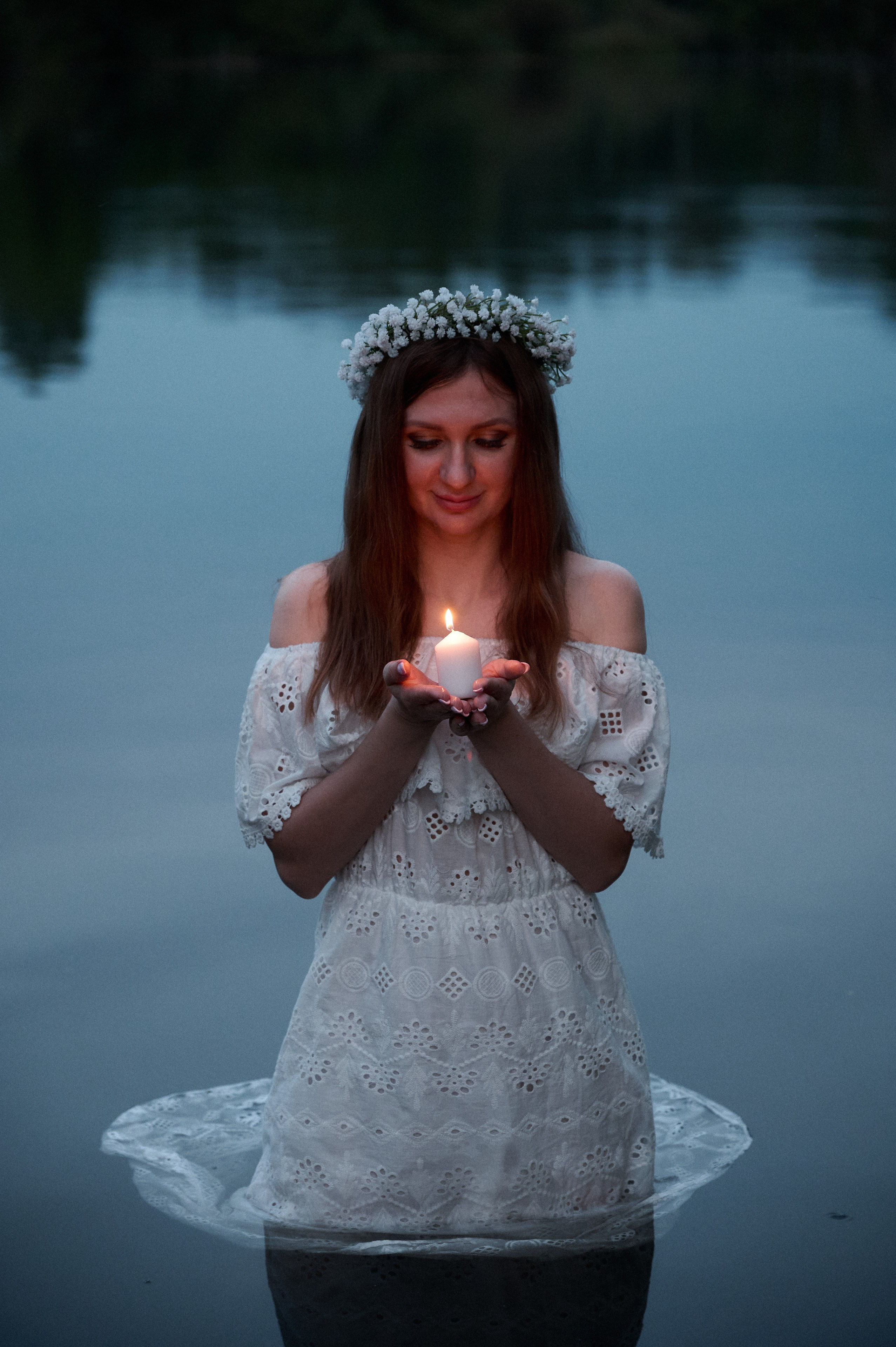 Евгения в белом платье стоит в воде, держит горящую свечу обеими руками.