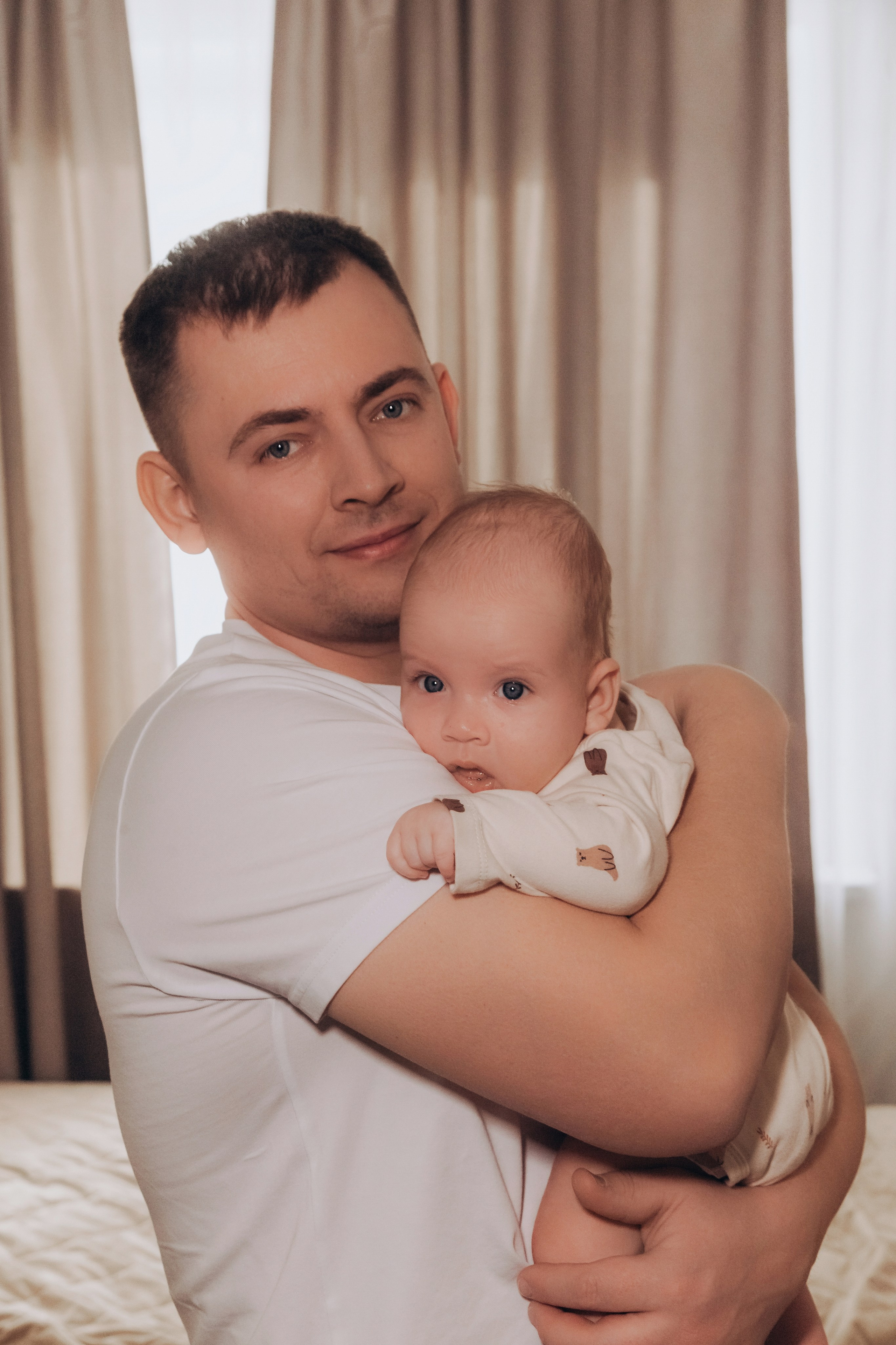 Папа стоит у кровати и держит младенца на руках, оба смотрят в камеру.
