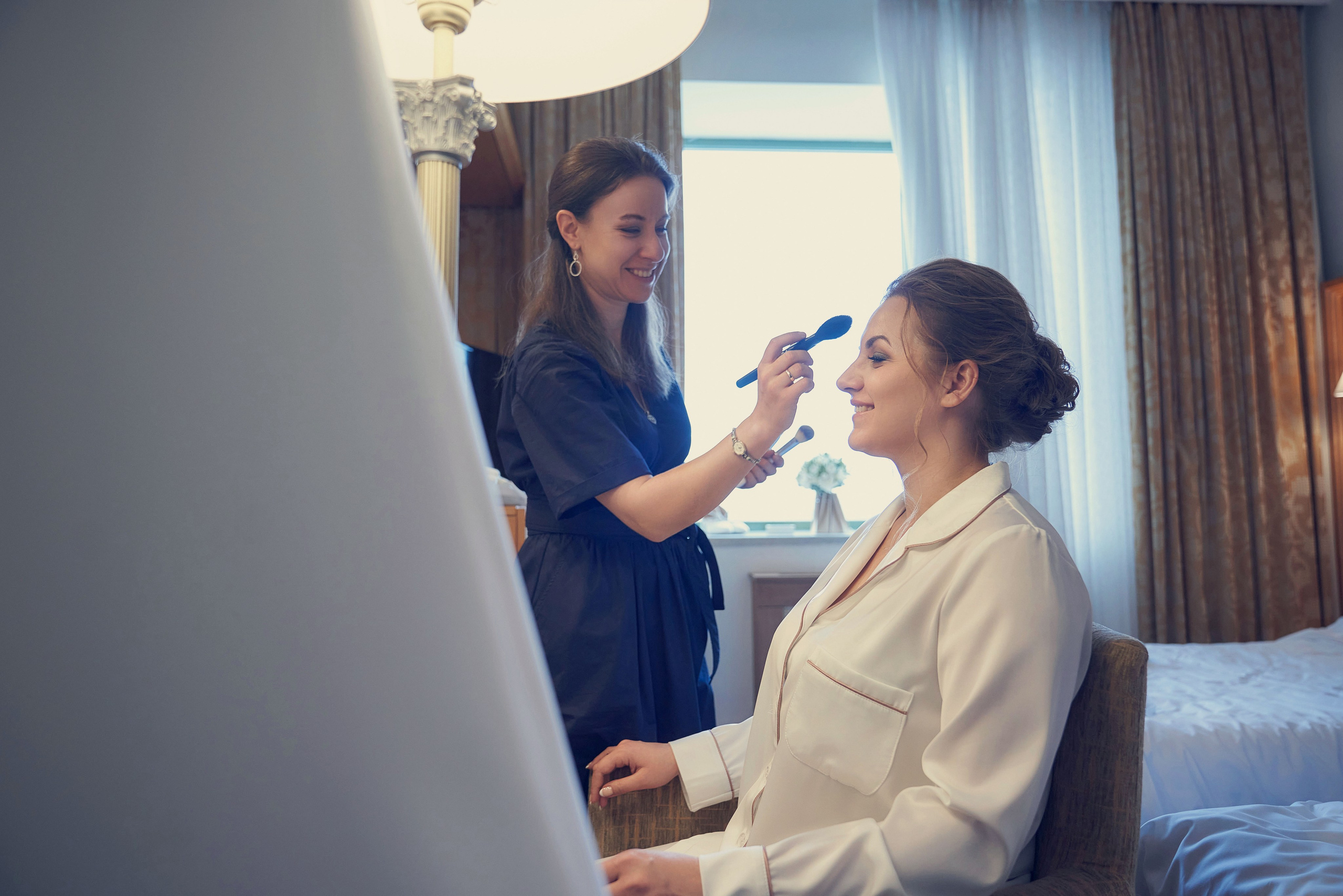 Визажист делает мейкап невесте в номере отеля на фоне свадебного платья и окна