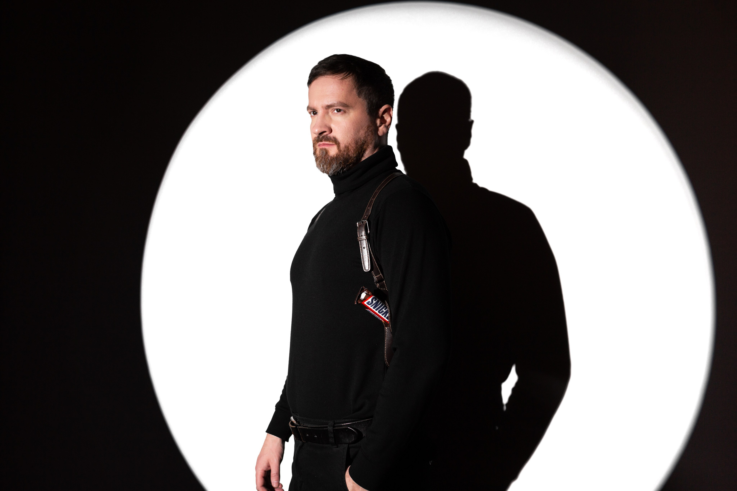 Брутальный мужчина с бородой и портупеей в белом круге света. Мужчина в черной водолазке. Мужчина со сникерсом.