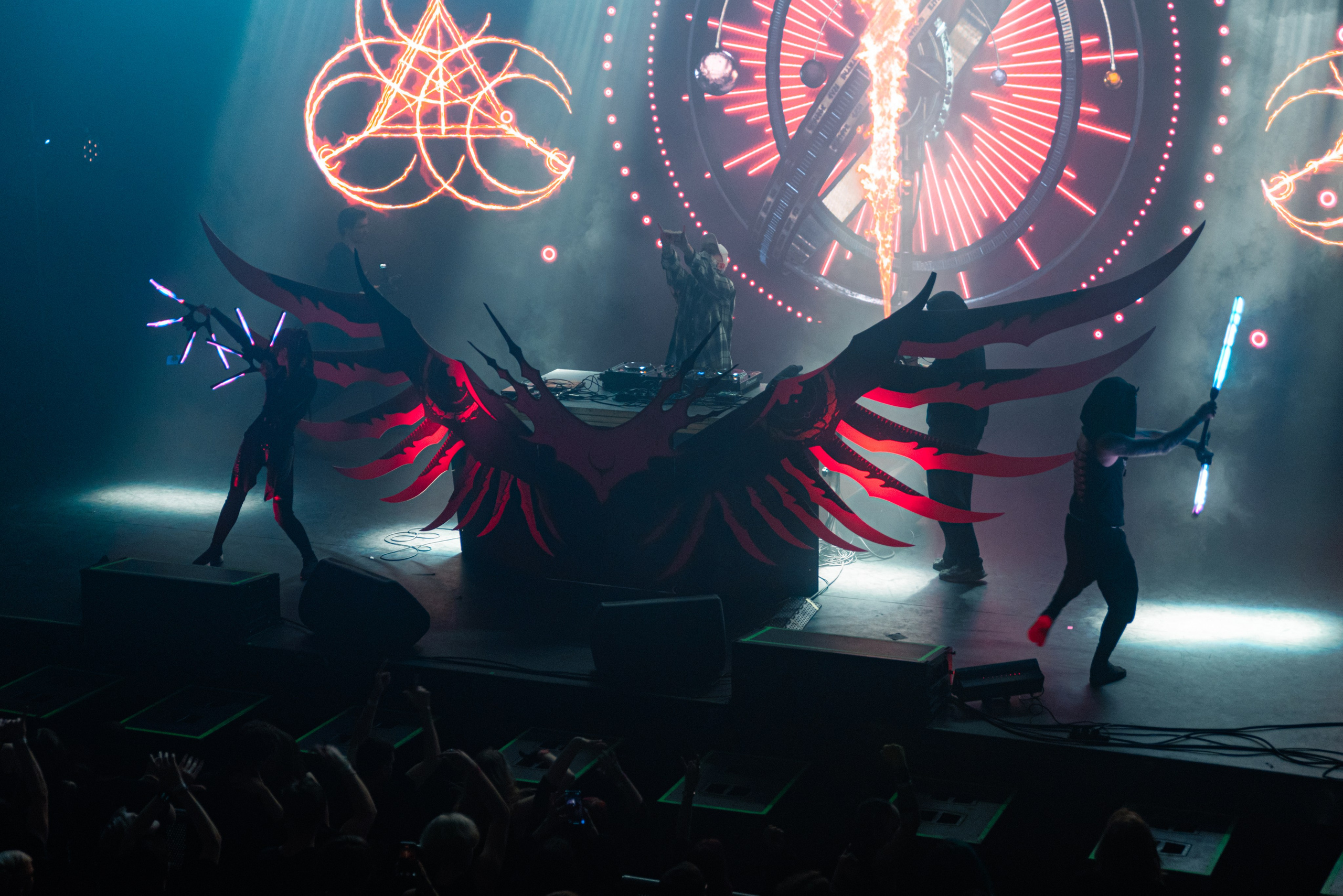 Сцена, посередине инсталляция с огромными крыльями и пульт диджея, по бокам два танцора с длинными светящимися палками