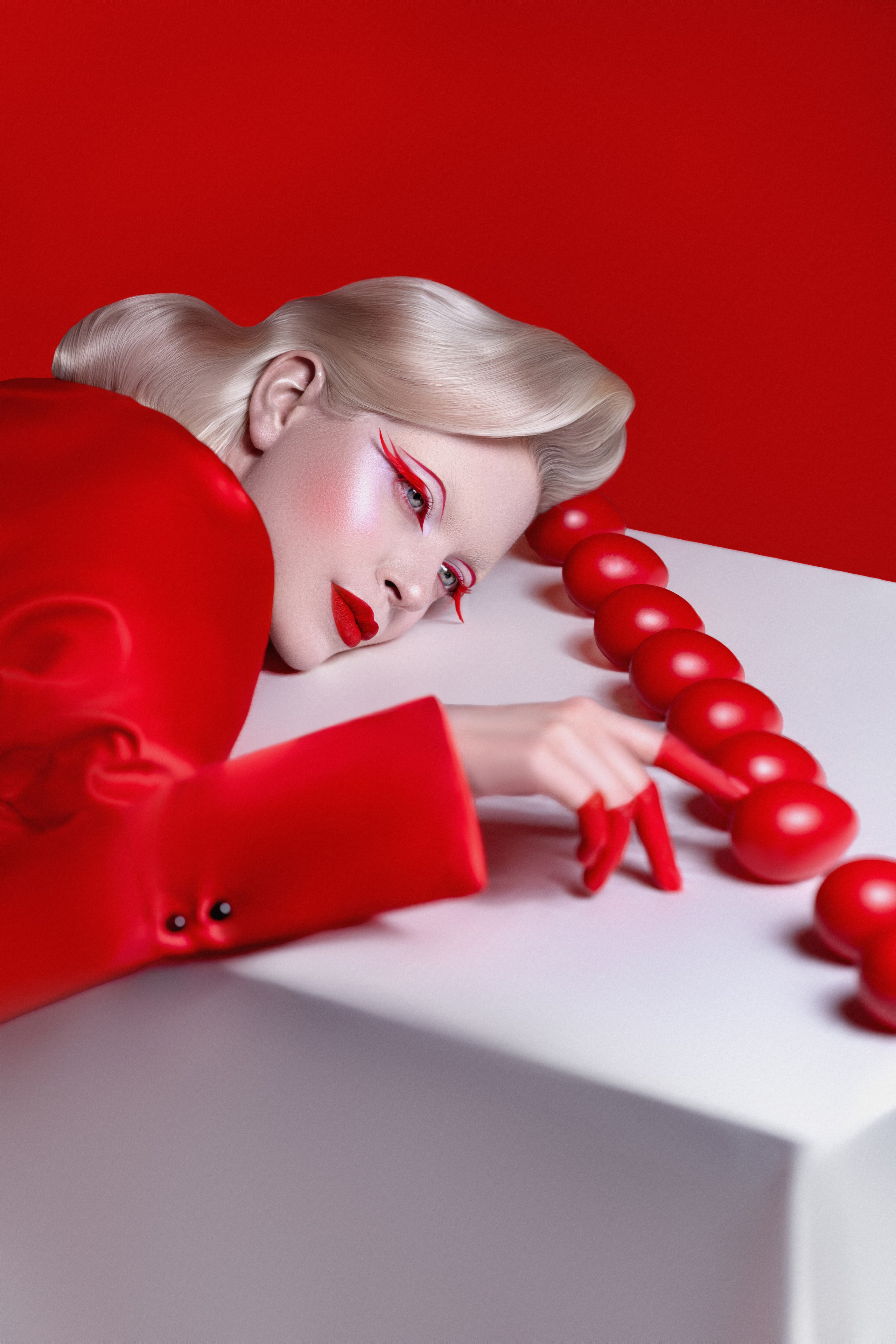 Блондинка с ретро-прической и выразительным красным макияжем в красном костюме лежит на белой поверхности, указывая на ряд красных шаров. Современная fashion-фотография, минимализм, авангард, beauty-портрет, креативный мейкап, арт-стиль, красный total look, editorial фотосессия, Instagram-контент, глянцевый стиль