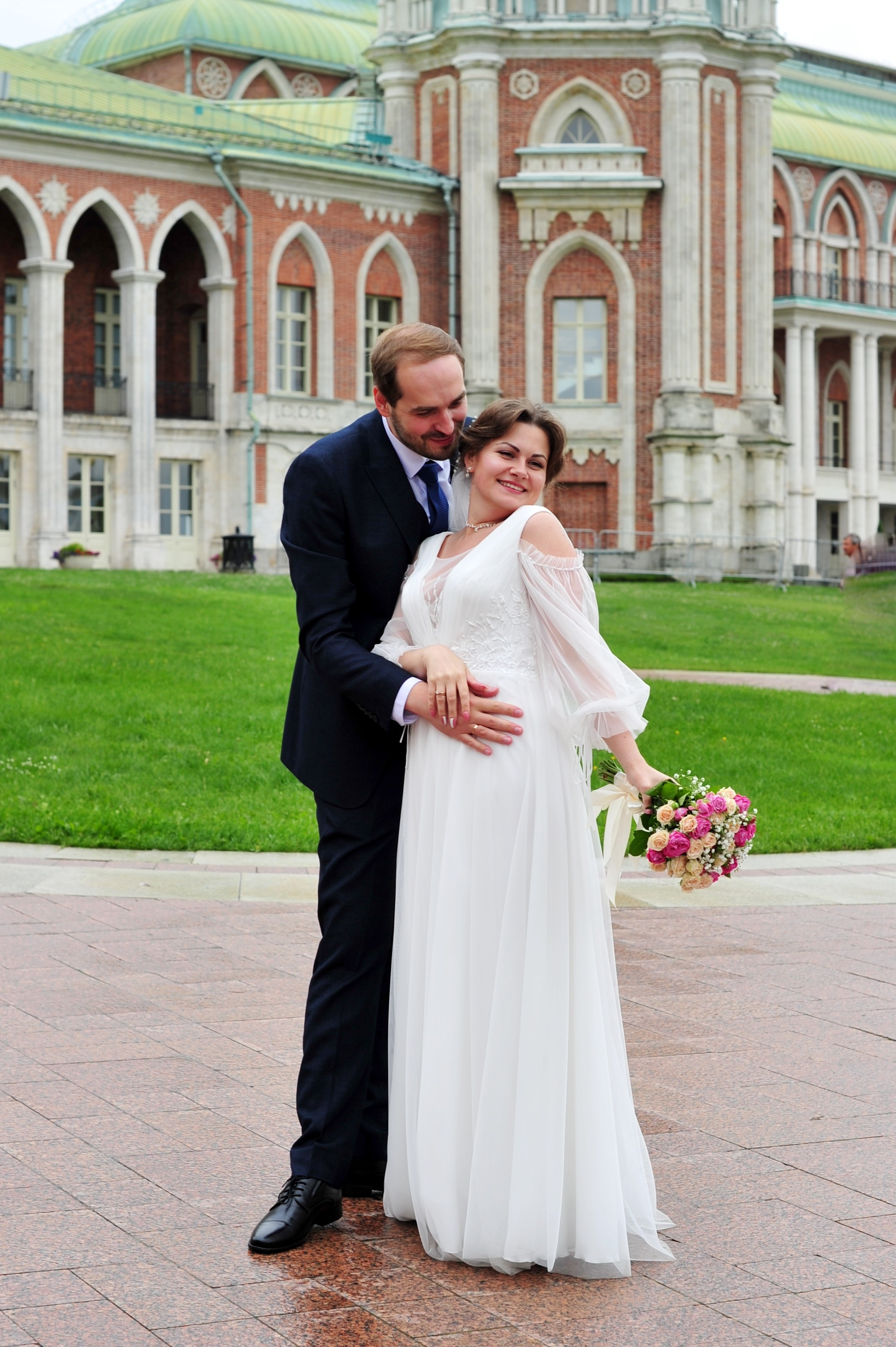 Свадьба в Царицыно пасмурным летним днем, новобрачные в полный рост на фоне дворца из красного кирпича 
