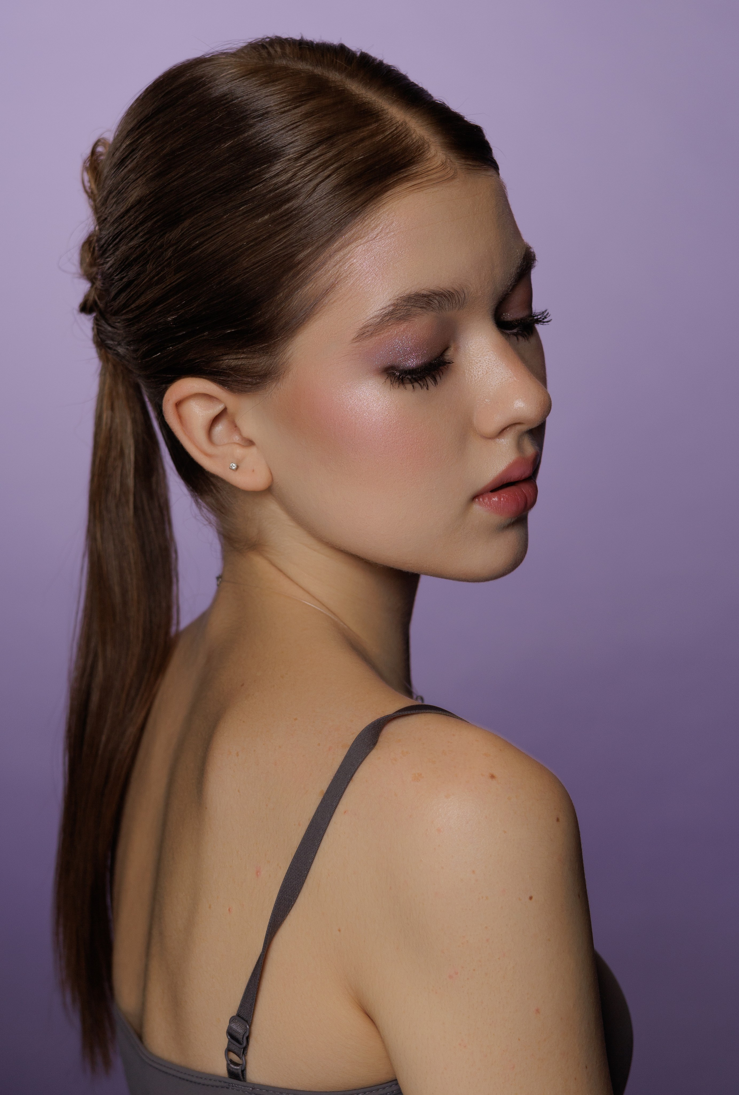 Портрет молодой женщины с гладкими каштановыми волосами, собранными в низкий хвост, на нежно-фиолетовом фоне. 