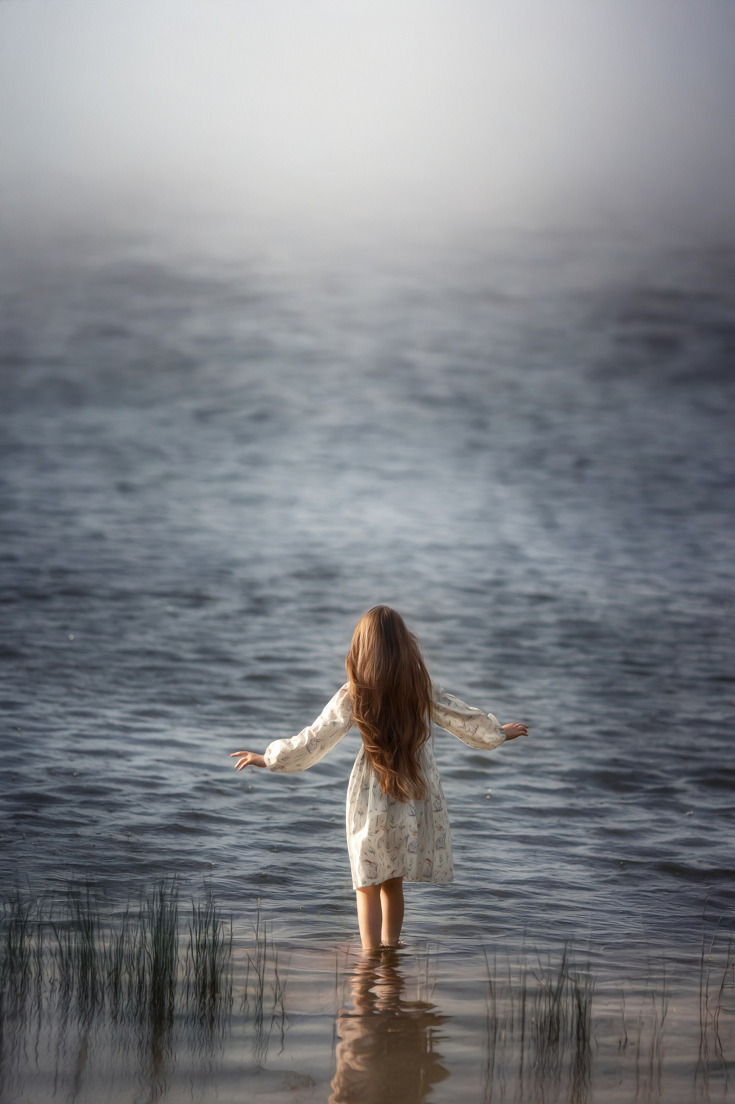 Девочка с длинными волосами стоит по колено в воде, раскинув руки, глядя в сторону тумана над озером.