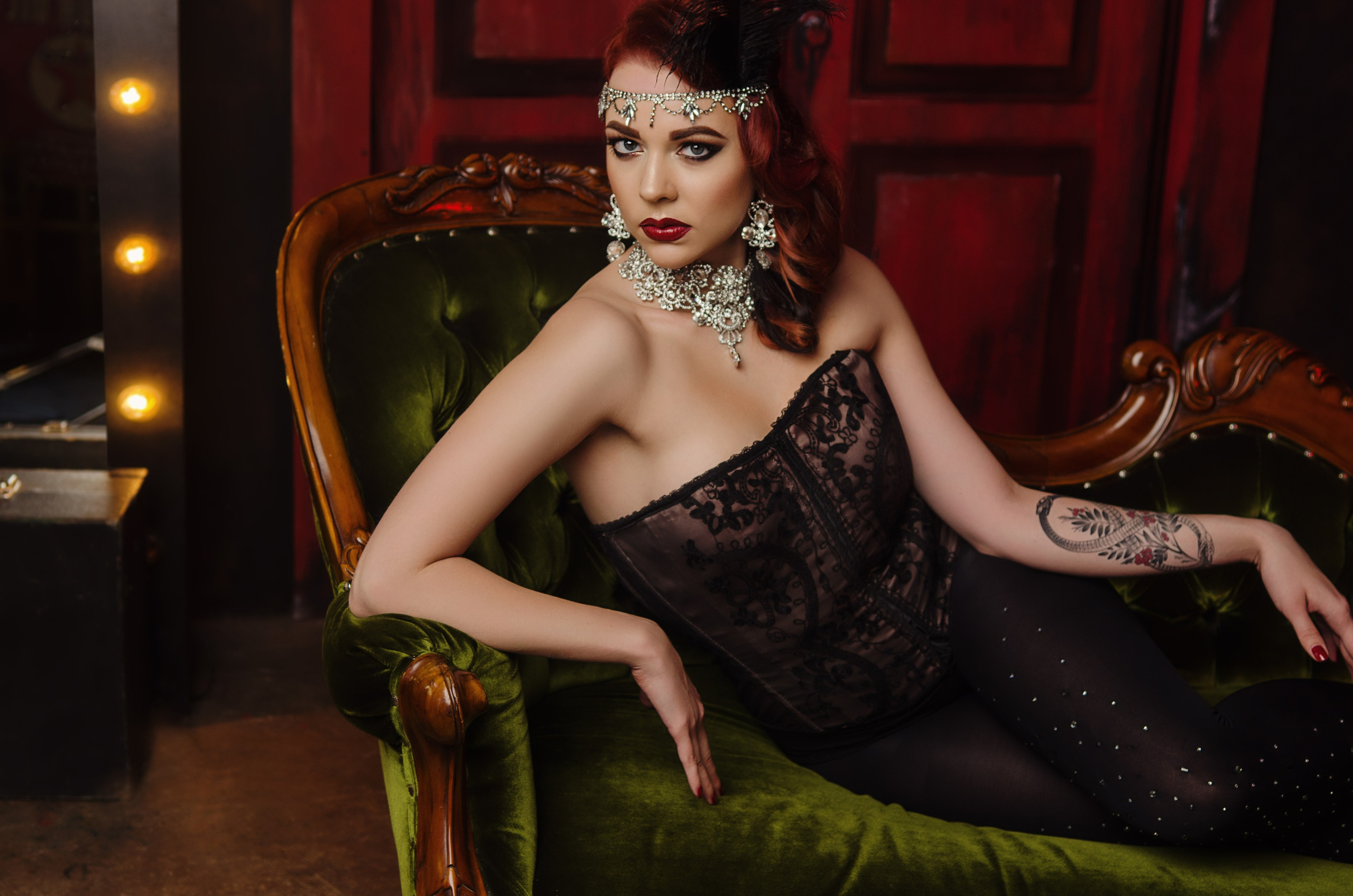 Девушка в стиле бурлеск с ярким макияжем и пором на голове сидит на зеленом бархатном диване