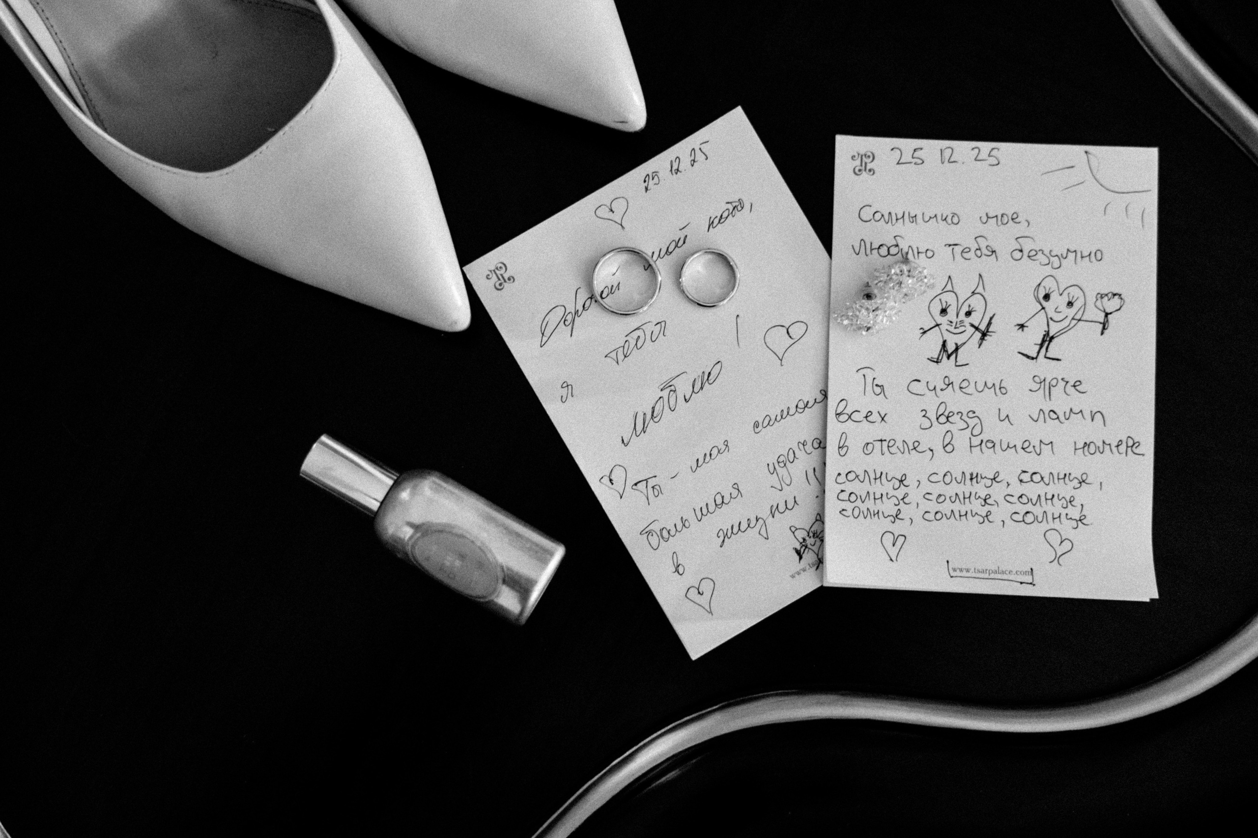Черно-белое фото двух писем, свадебных колец и маленькой бутылочки на темном фоне.