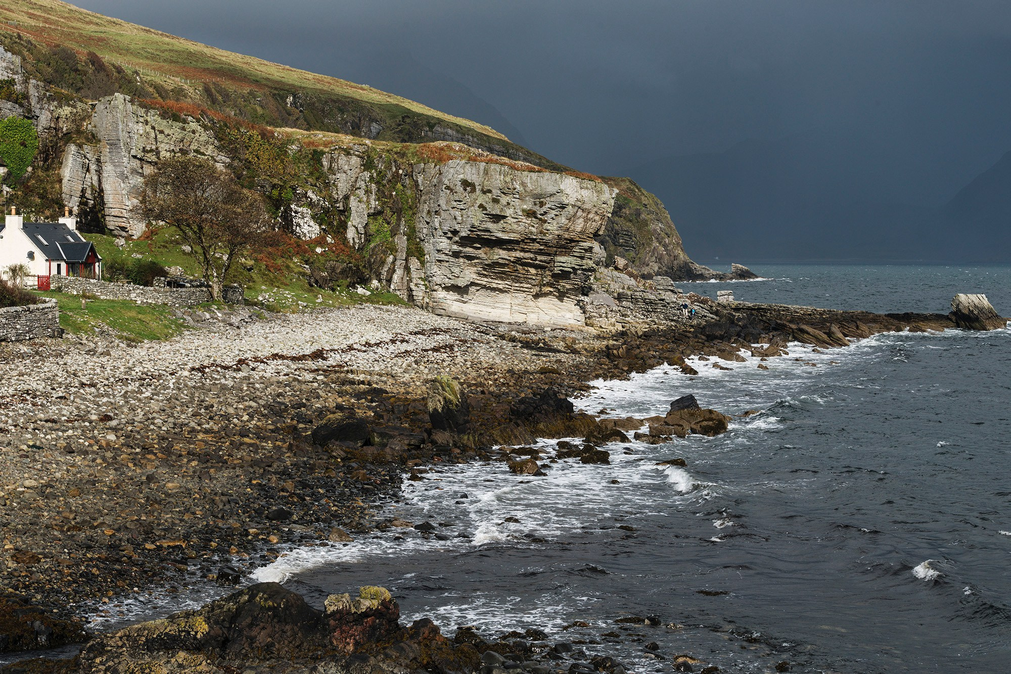 Осеннее фото пляжа Элгол на острове Скай в Шотландии: скалистый берег и темные волны океана, контрастирующие с ярко освещенными золотисто-зелеными склонами холмов и домом на побережье, драматичное небо.