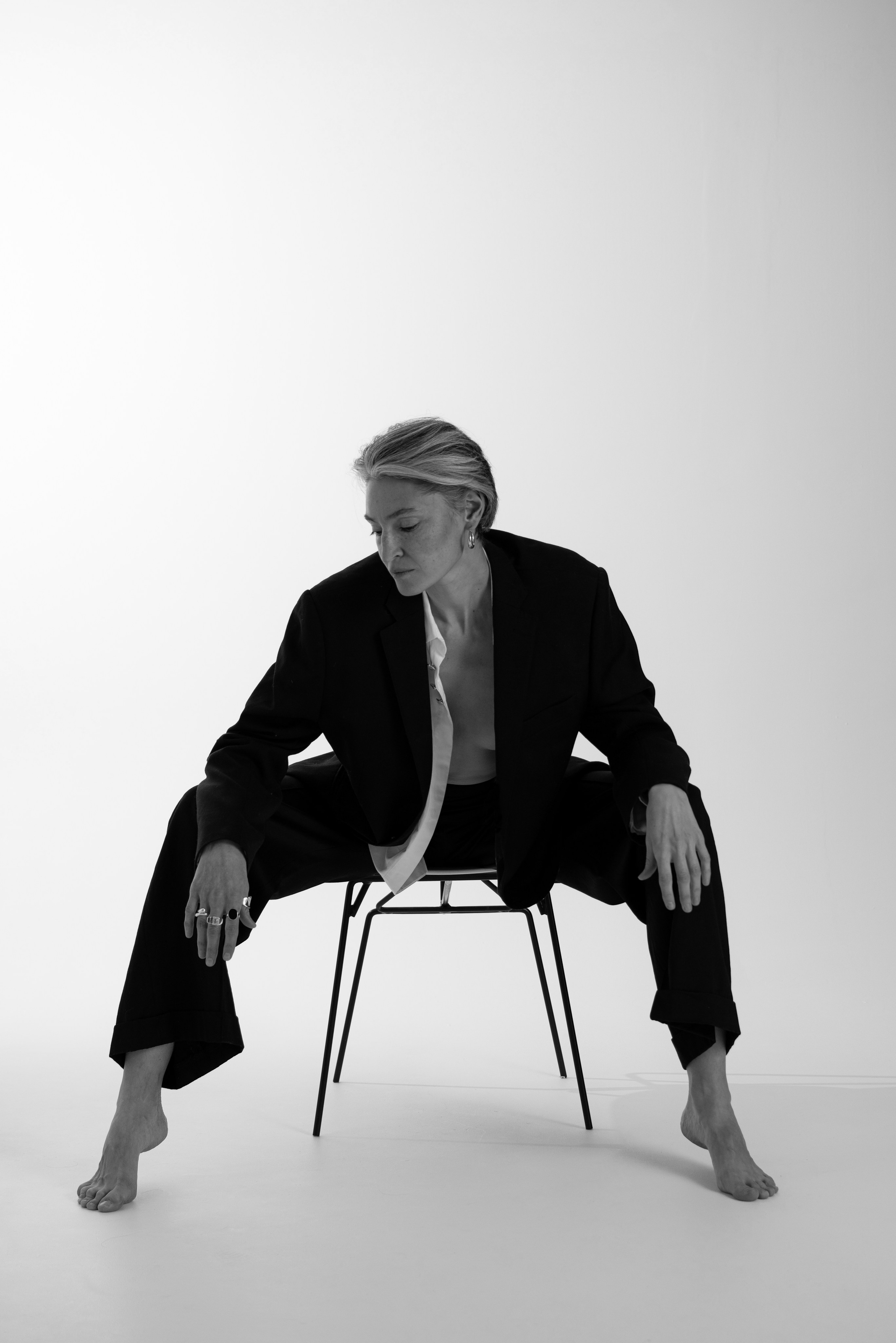 Модель в чёрном костюме сидит на стуле в минималистичной студии, позируя в расслабленной позе. Съёмка для бренда одежды.