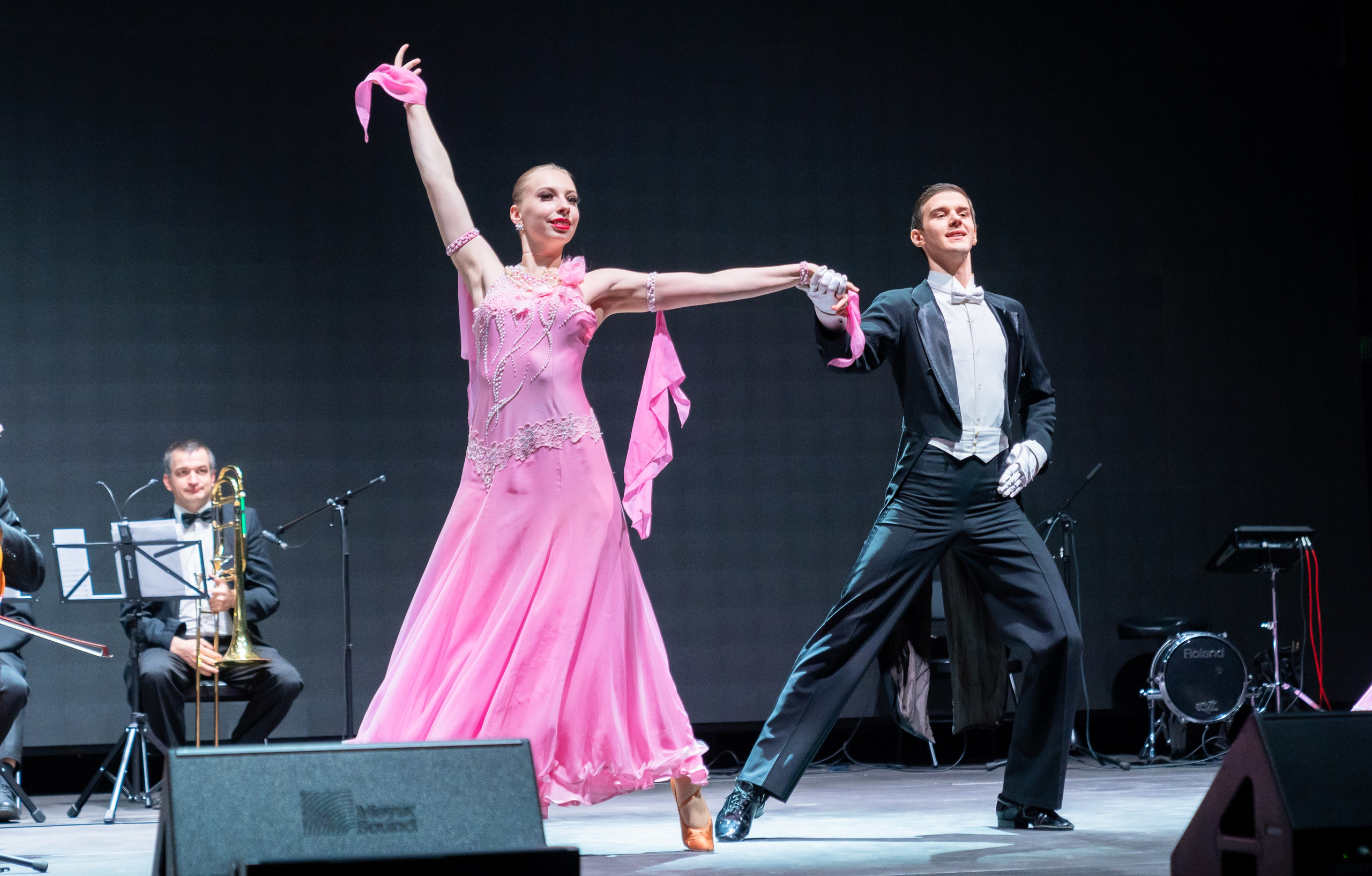 Танцоры в розовых костюмах: женщина в развевающемся розовом платье и мужчина в чёрном фраке исполняют бальный танец под аккомпанемент оркестра. День местного самоуправления СПб. Фото Евгения Мальцева.