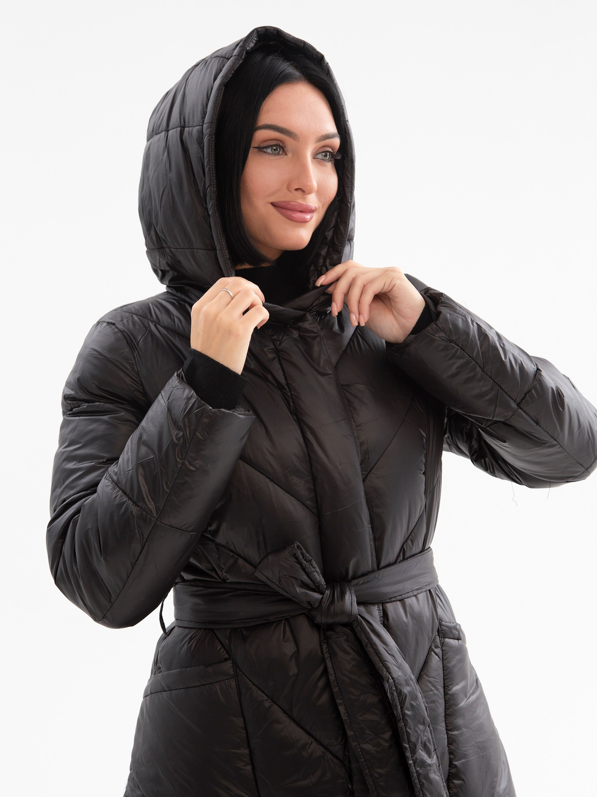 Девушка брюнетка с зелеными глазами в черном пуховике с капюшоном стоит на белом фоне