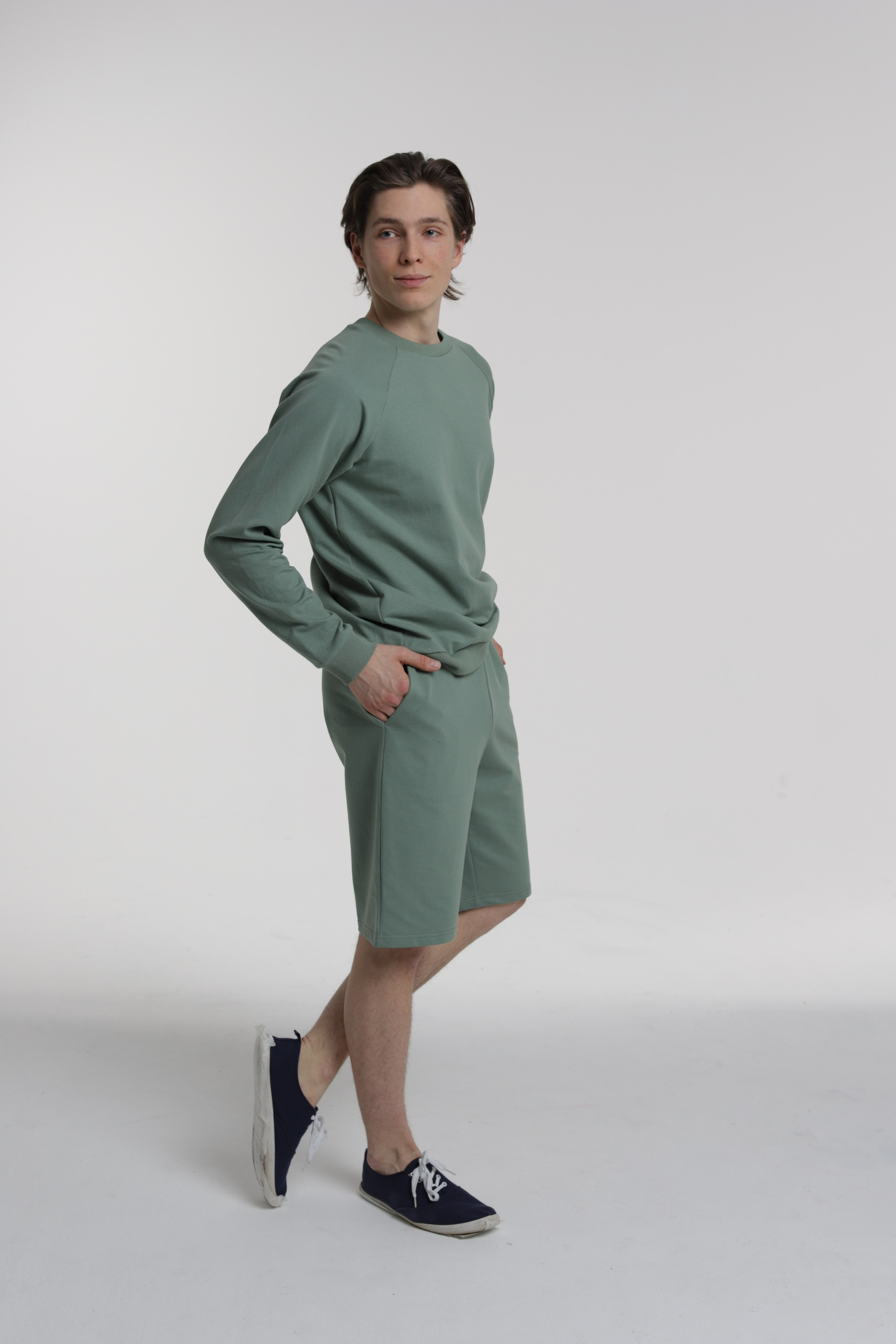 Молодой человек в зеленом лонгсливе и шортах позирует на сером фоне