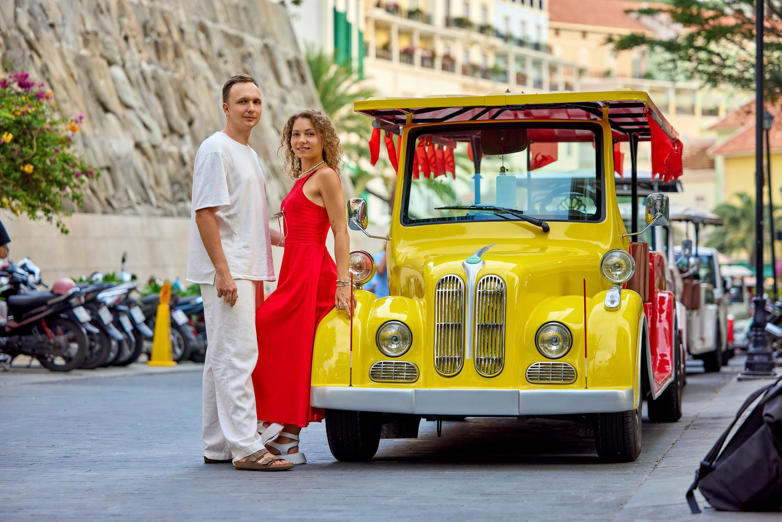 A smiling couple wearing white and red clothes standing next to a yellow classic-style electric car on a street in Sunset Town, Phu Quoc, Vietnam.