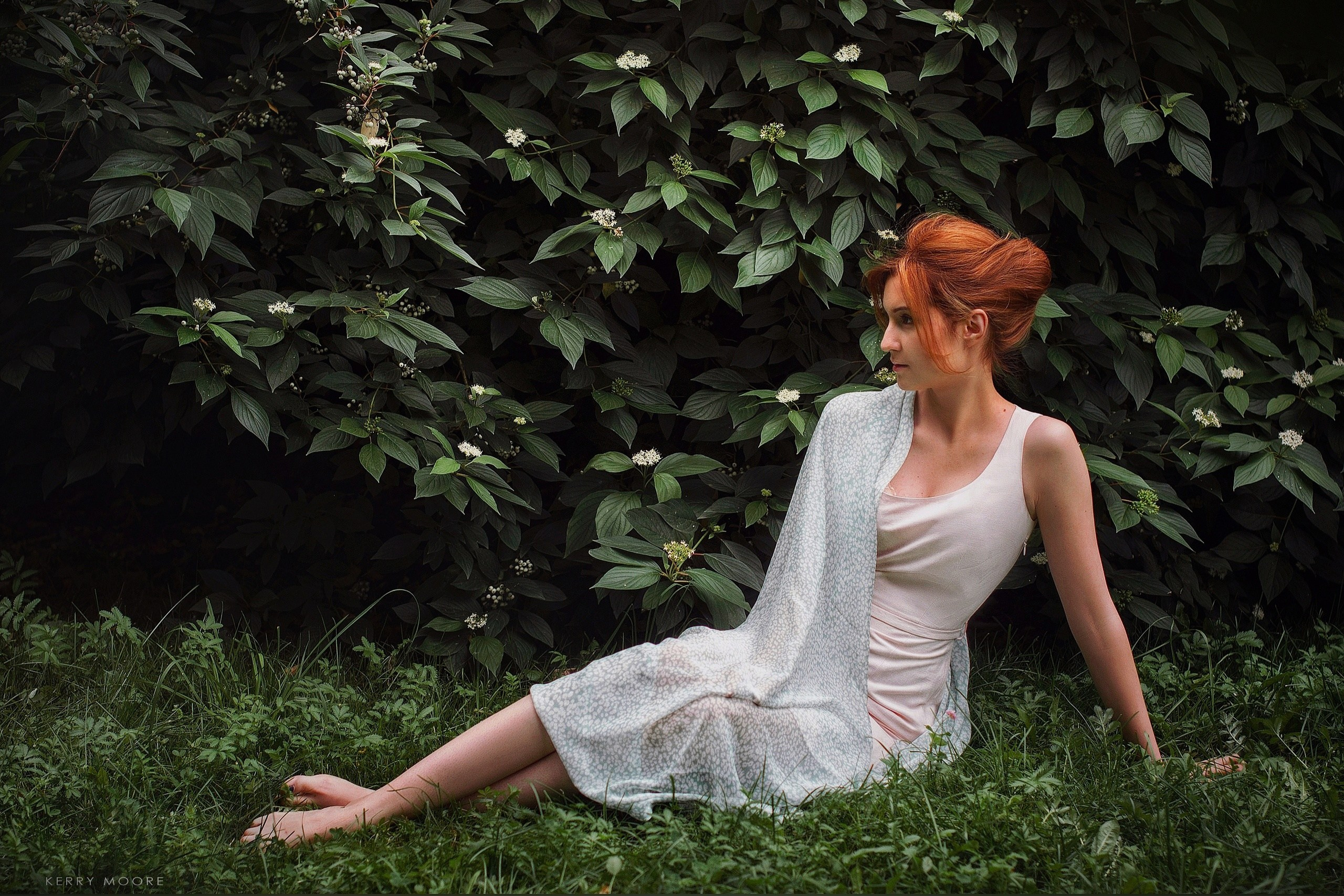 Девушка с рыжими волосами и в светлом платье изящно сидит на траве 