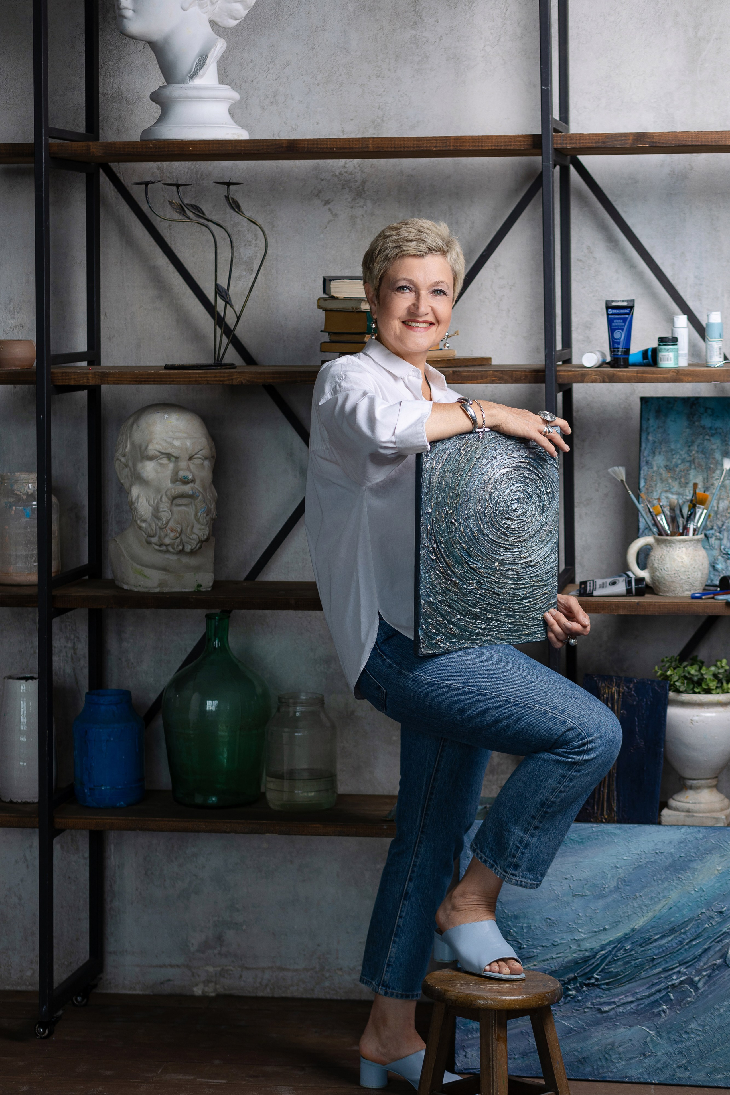 Художница стоит у стеллажа, правую ногу поставив на табурет.