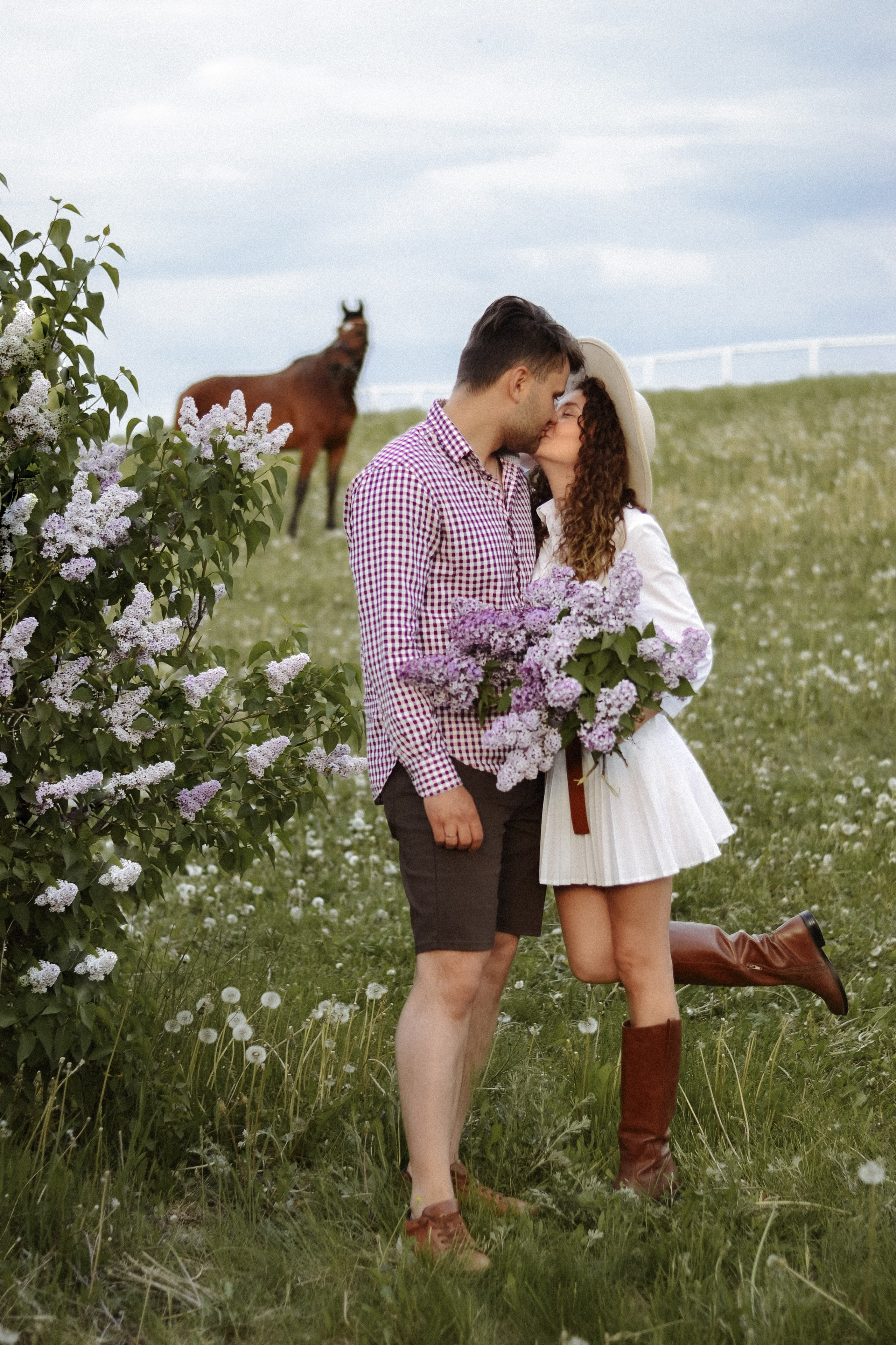 На фотографии изображены два человека, которые обнимаются на фоне цветущего поля. Мужчина одет в клетчатую рубашку и коричневые шорты, а женщина носит белое платье с цветочным узором и широкополую шляпу. В руках у нее большой букет фиолетовых цветов. На заднем плане видна лошадь, стоящая на траве. Эта сцена создает ощущение летнего дня, проведенного на природе.