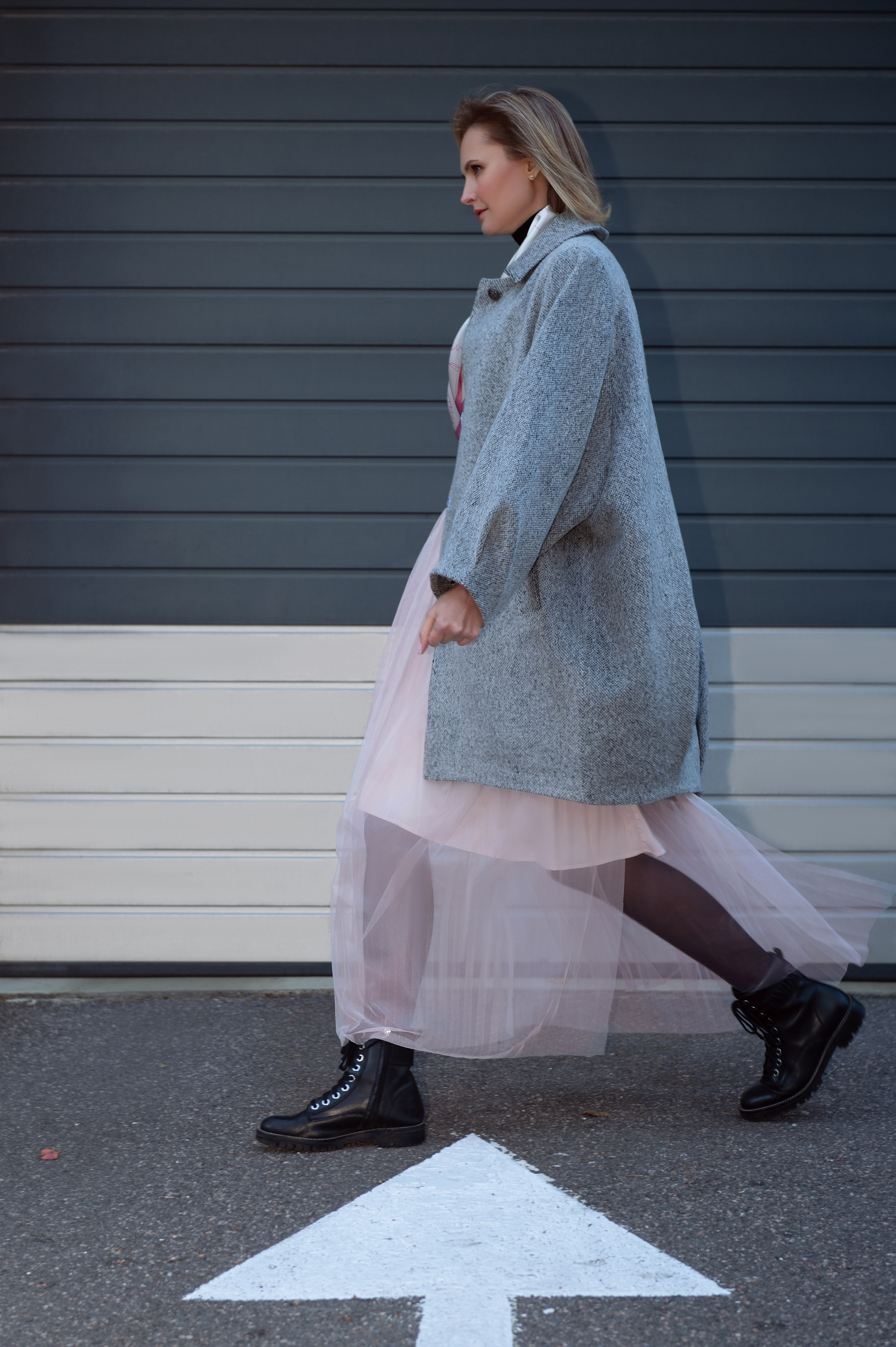 Женщина идет вдоль серой стены. На ней серое пальто-кокон, розовая юбка из фатина и черные грубые ботинки. Уличная контент-съемка для бутика. Съемка в динамике.