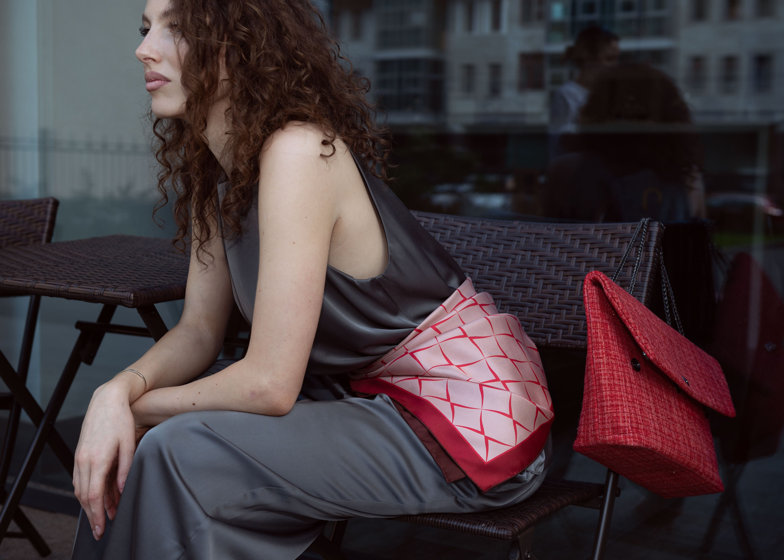 Красивая девушка в сером брючном костюме и красном шелковом дизайнерском платке на поясе сидит за столиком в уличном кафе