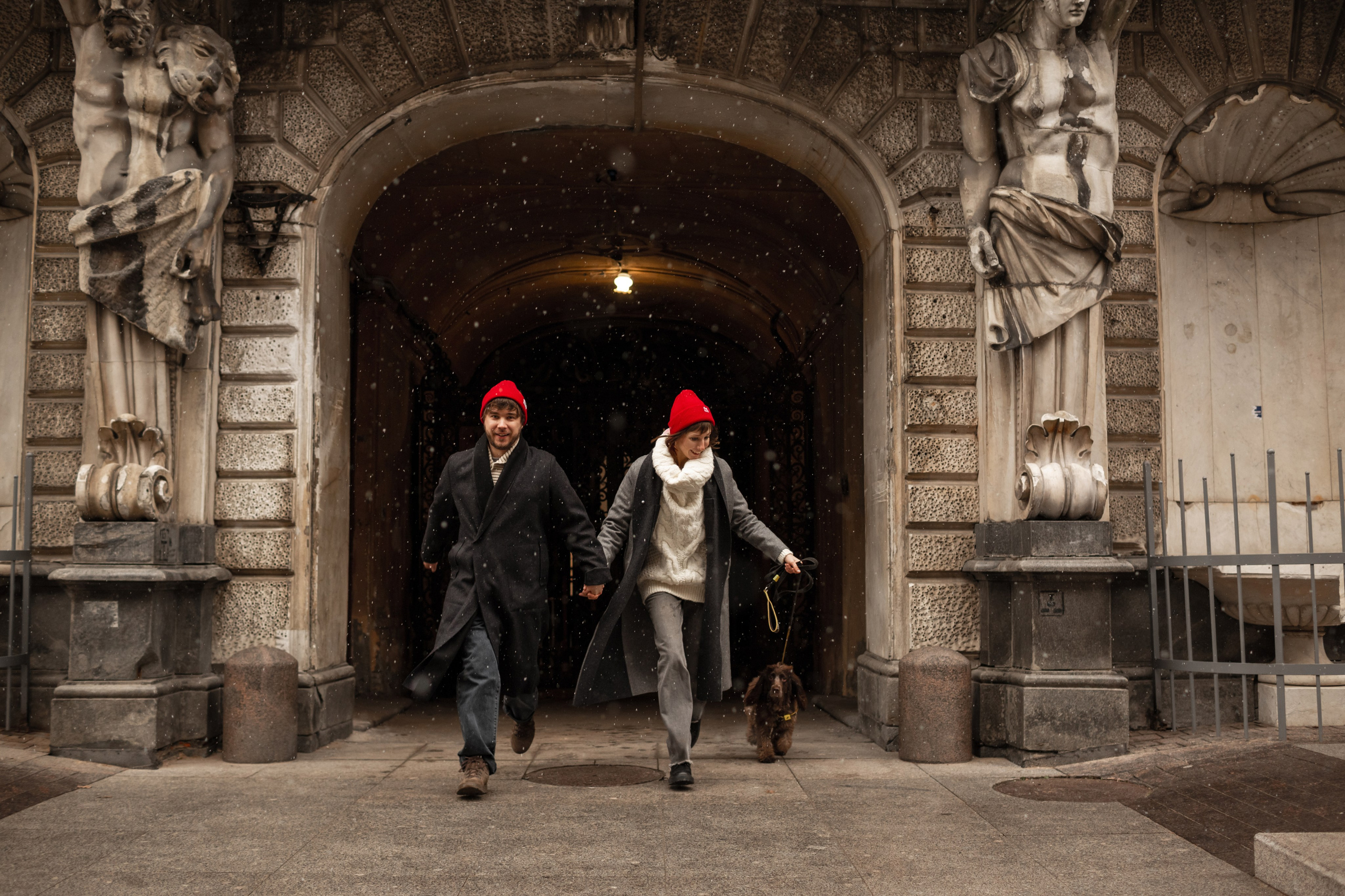 Молодая пара в парных красных шапках Kusto бегают с собакой на фотосессии.