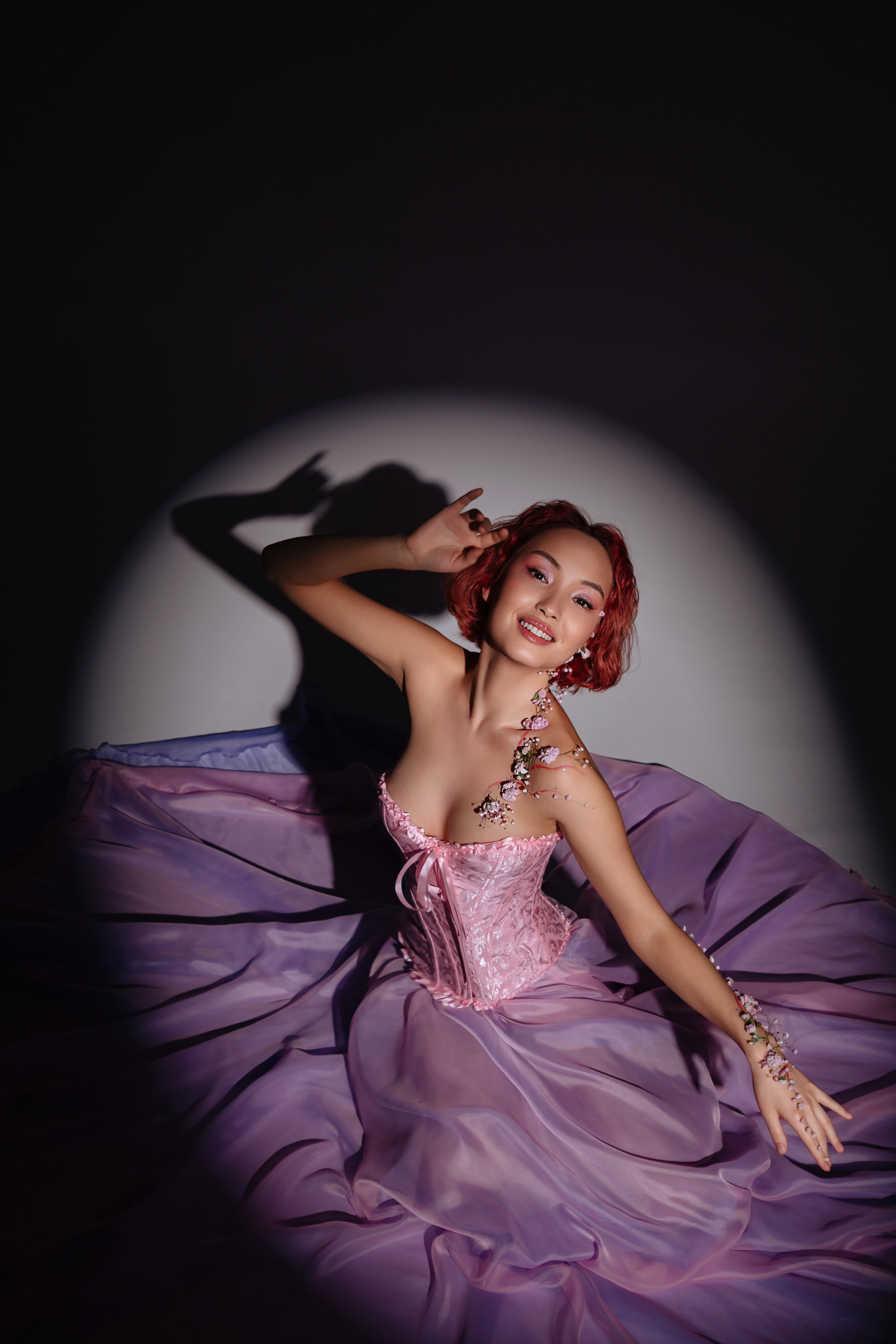 Чувственная сцена, где юная азиатка в живых цветочных украшениях погружена в круг мягкого света, ее задумчивый взгляд предвещает приход весны.