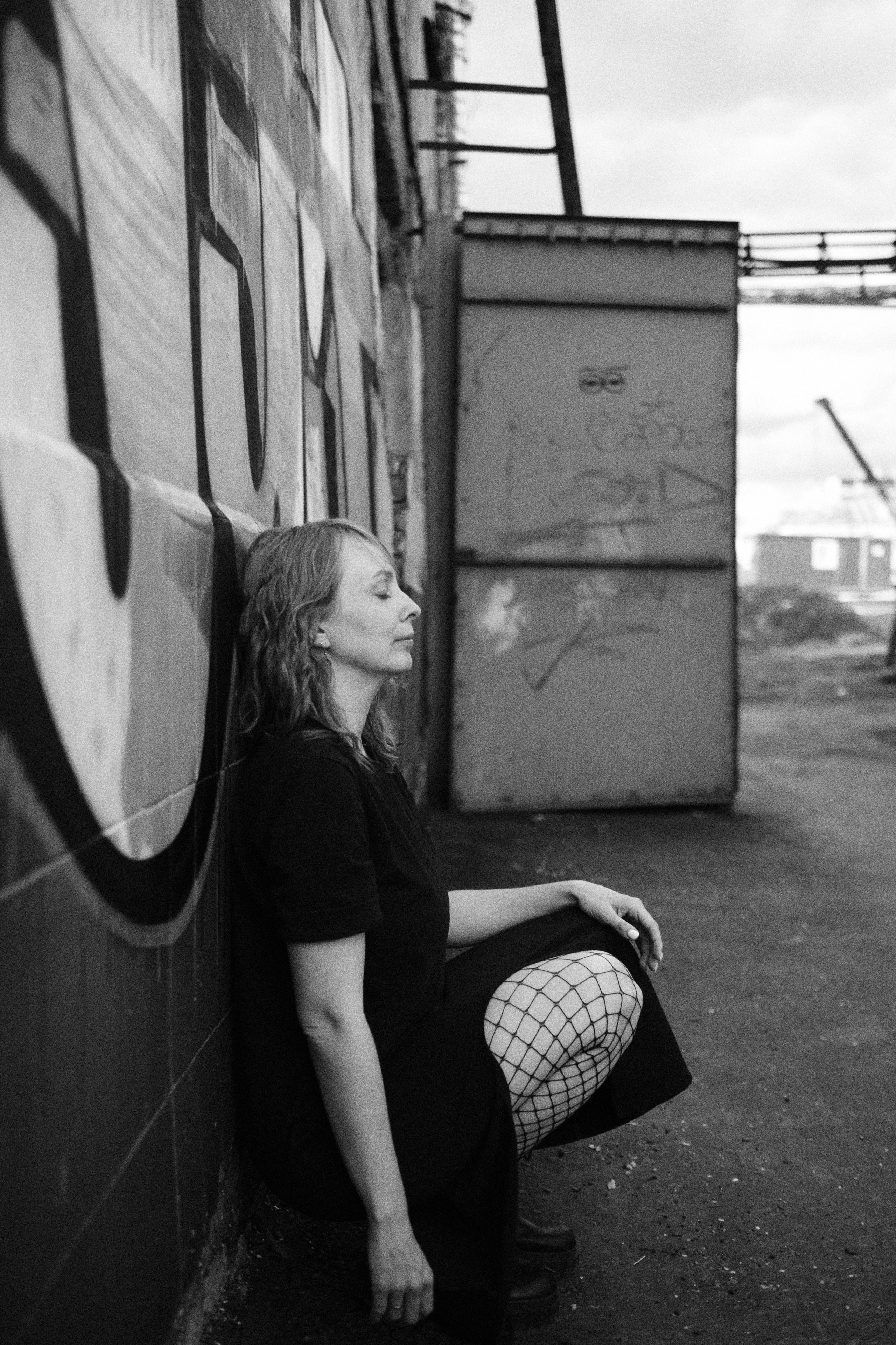 Женщина в черном платье сидит на корточках, облокатившись о стену