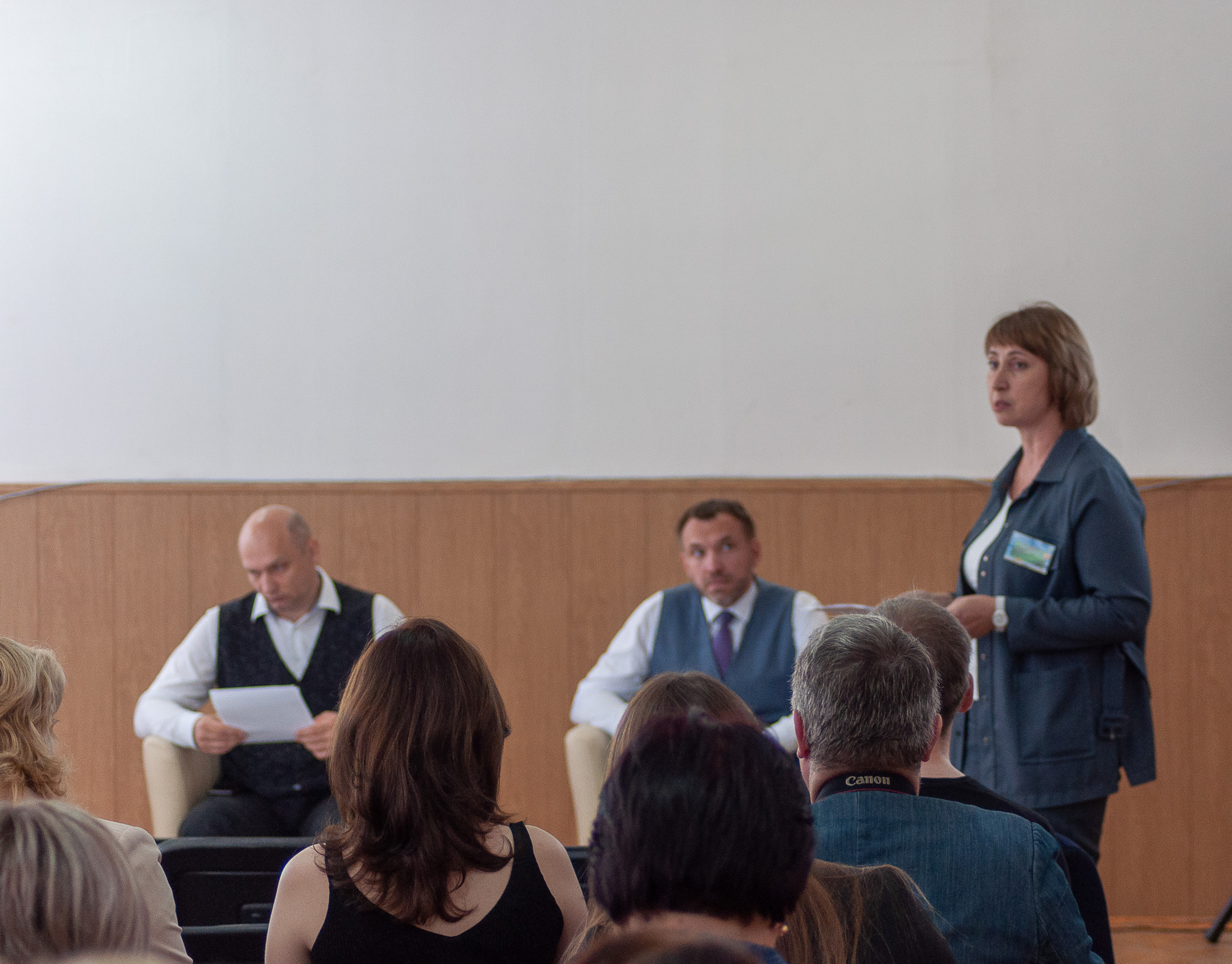 Женщина выступает с докладом перед аудиторией, мужчины на сцене слушают и читают материалы.