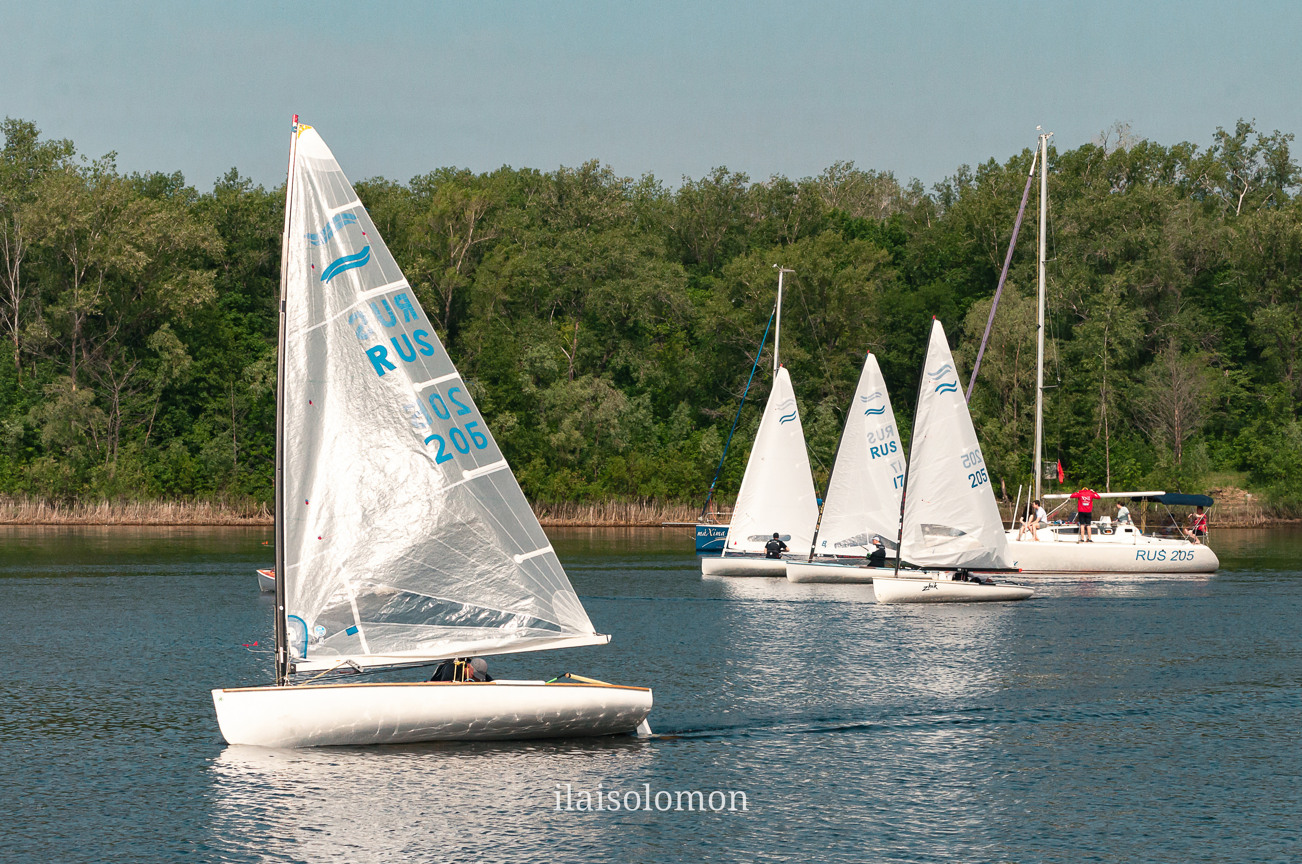 Парусная яхта набирает скорость, сзади три лодки и судейское судно на фоне берега.