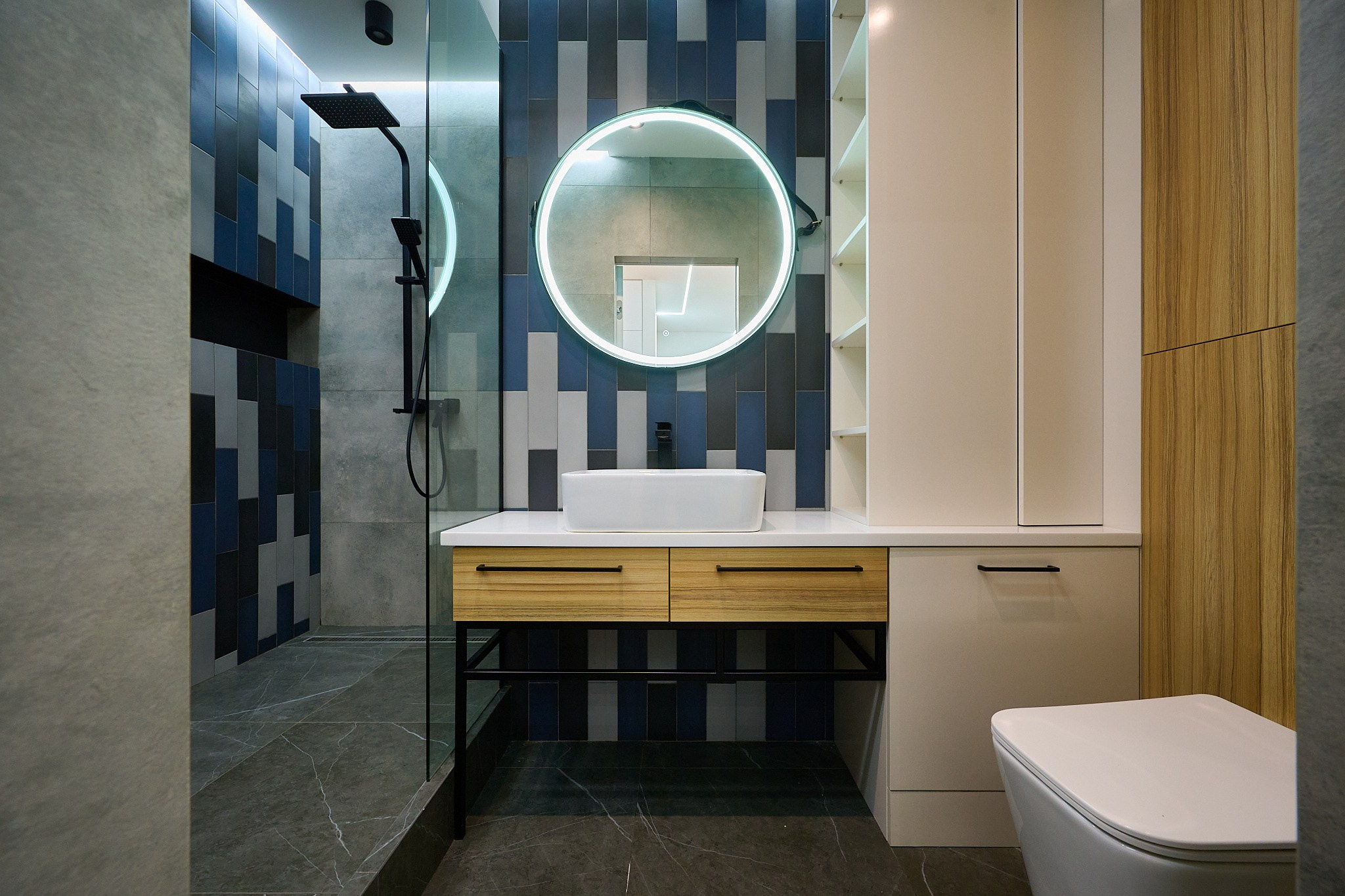 ванная комната с круглым светящем зеркалом с голубыми черными и белыми плитами на стенке и деревянными шкафами 