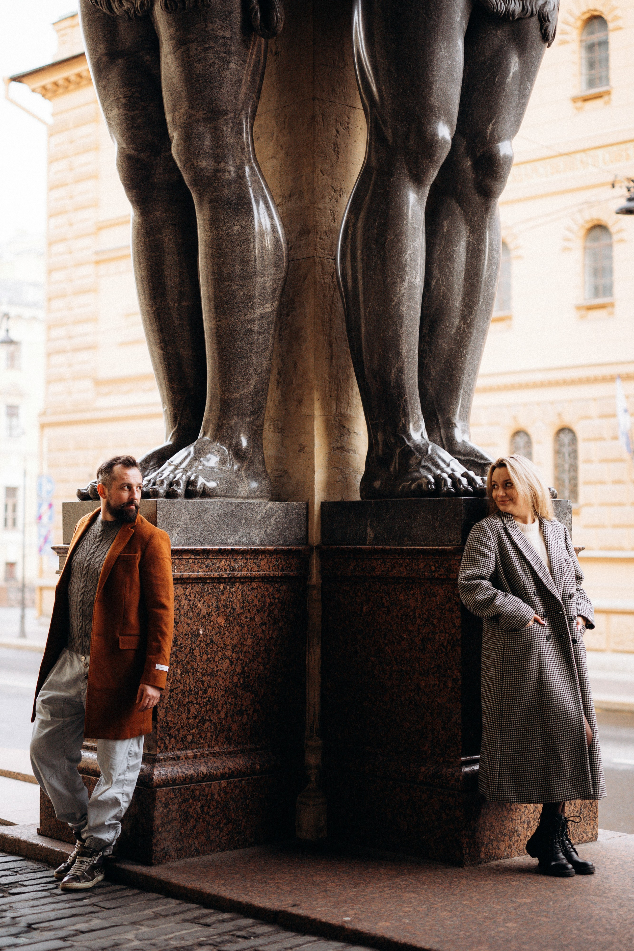 Лавстори Love Story в Санкт-Петербурге у Атлантов. Пара в коричневых пальто стоят рядом с ногами Атлантов