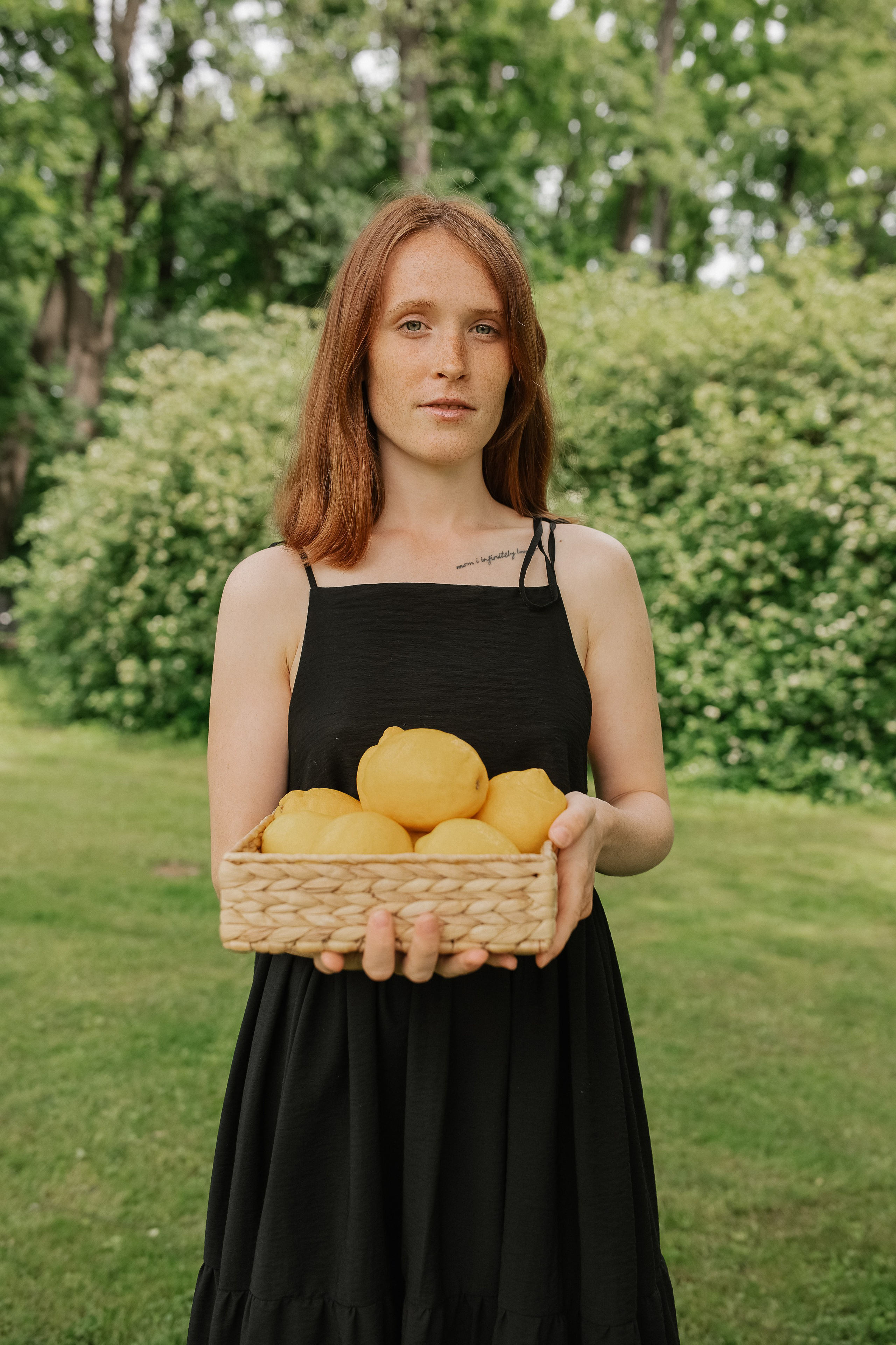 Рыжая девушка в черном платье держит в руках корзинку с лимонами