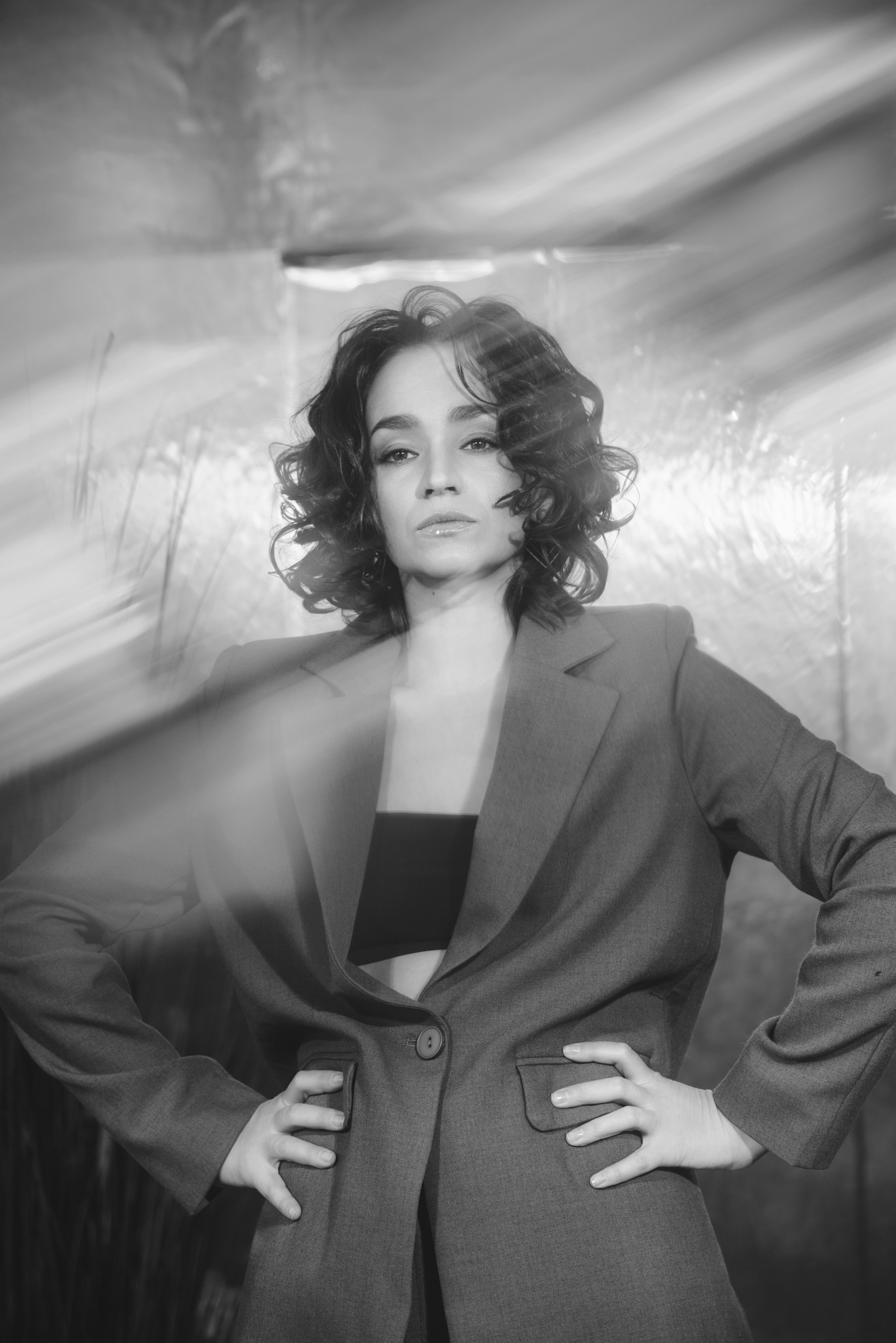 Портретное фото молодой девушки в студии. Девушка сидит на фоне нейтрального цвета, ее лицо освещено мягким светом. Она смотрит прямо в камеру с легкой улыбкой. На девушке стильная одежда, подчеркивающая современный модный стиль. Фотография выполнена в высоком разрешении, с акцентом на деталях