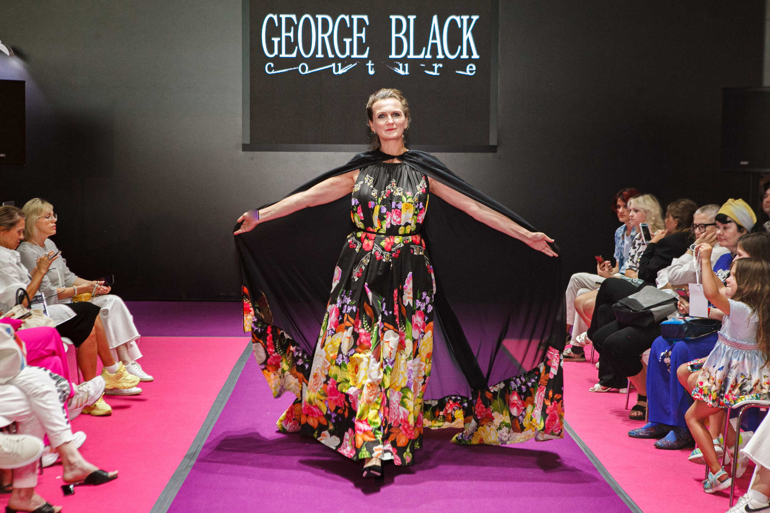  "Модели в ярких нарядах проходят по подиуму во время показа мод, освещенные яркими софитами и окруженные восторженными зрителями."