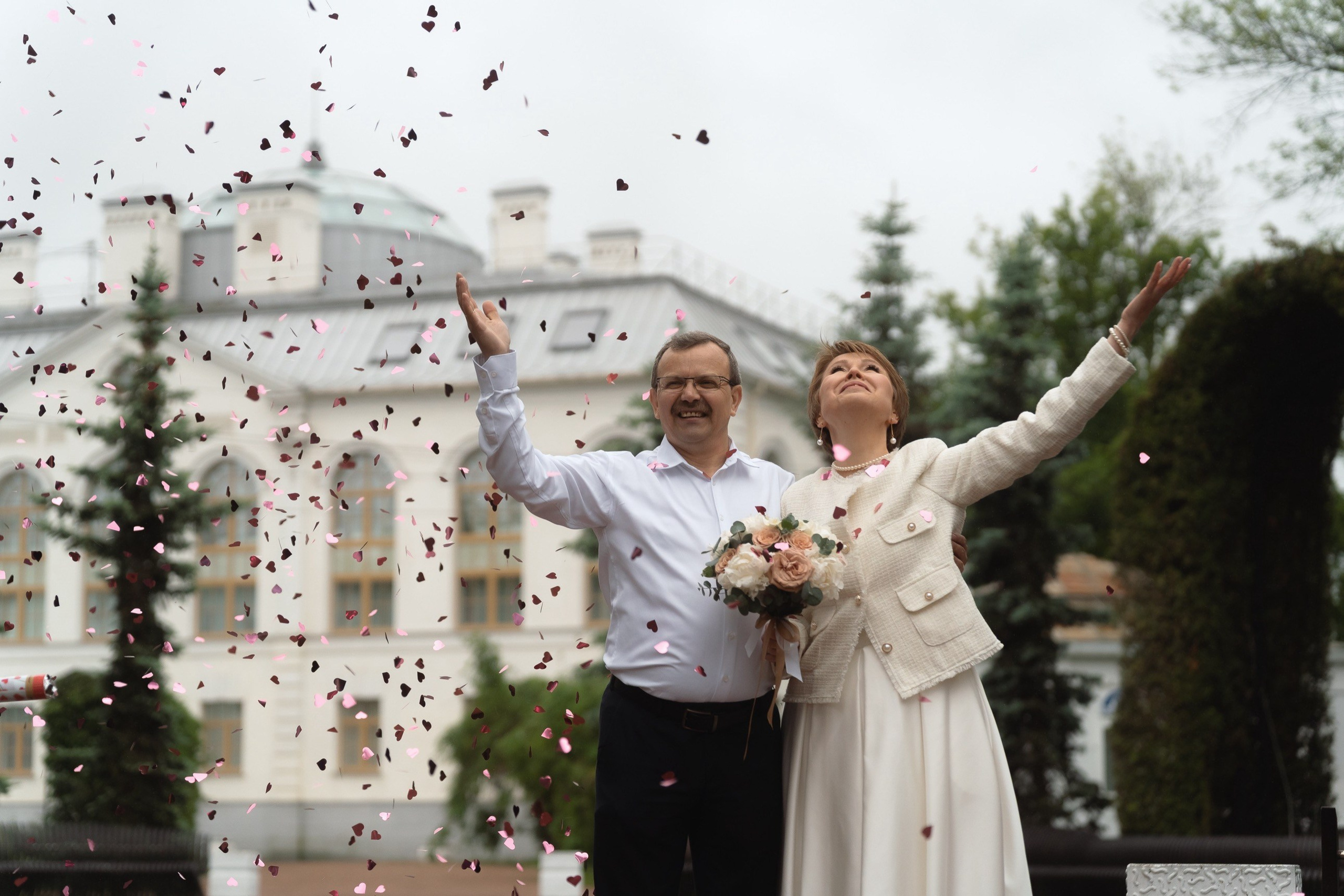 Молодожёны поднимают букеты и руки, под дождём лепестков у Дворца бракосочетаний в Санкт-Петербурге