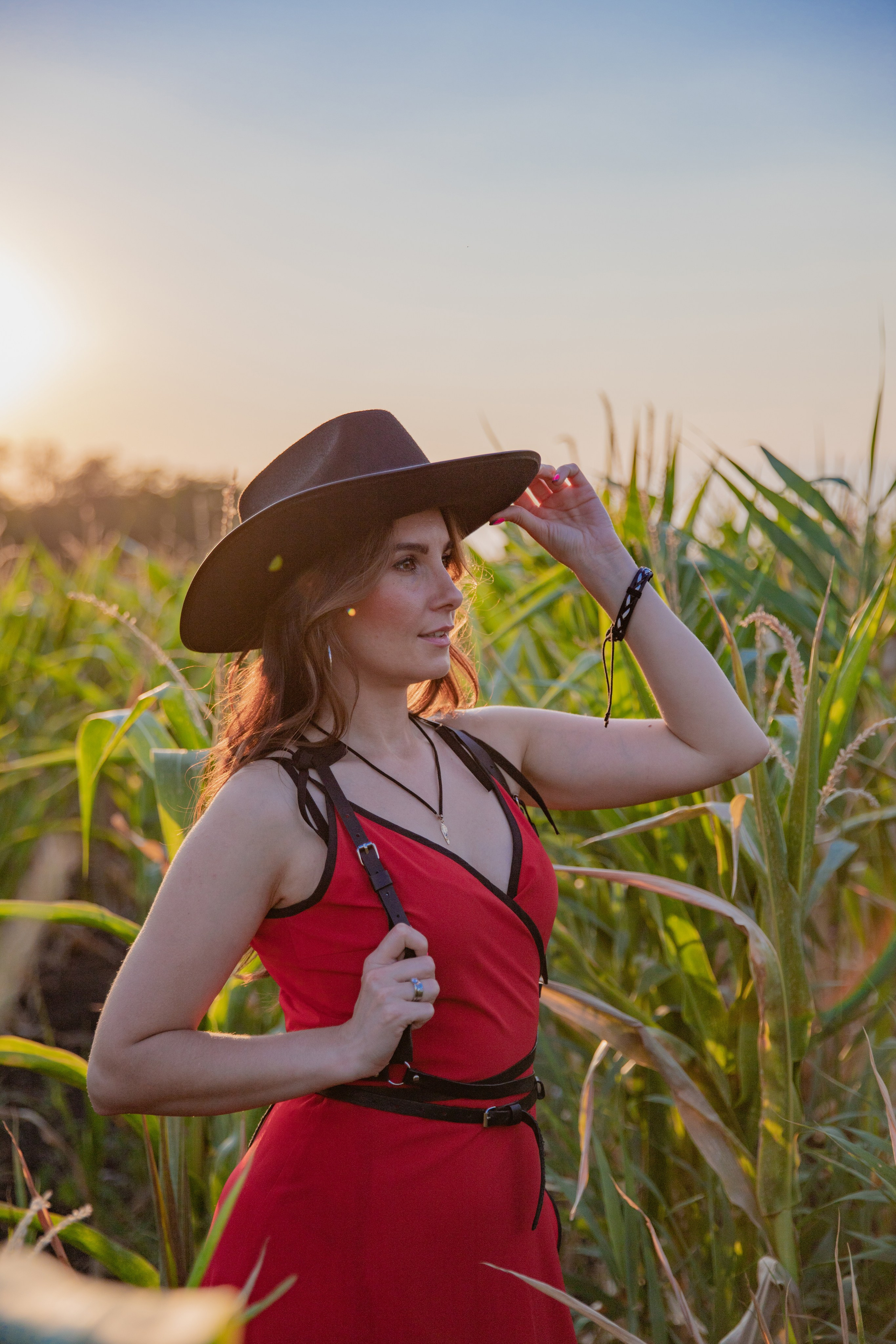 Портрет девушки в шляпе и в красном платье, улыбающейся на фоне кукурузного поля, с золотистыми лучами заката.