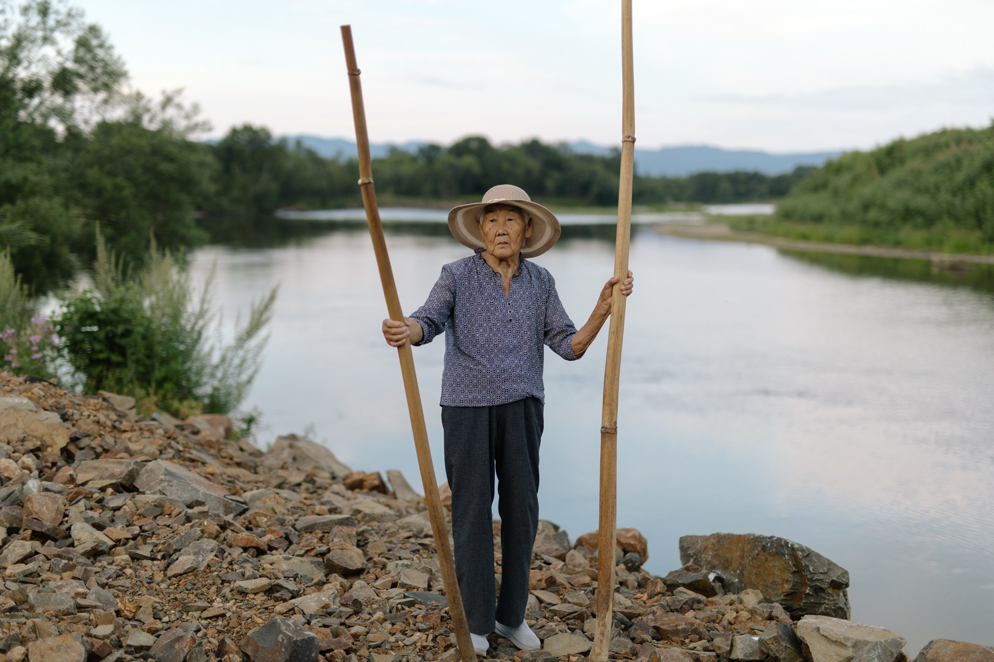 Efrosinya Shkalygina (Nivkh) on the Tym River bank in Chir-Unvd village. She is holding the ancient Nivkh kalni musical instruments in her hands. Kalni is made from a dry bear pipe, a hollow plant one and a half to two meters high, leaves are cut off. The performer blows into it, vibrating ones voice. Previously, the instrument served as a kind of sound beacon: if there was fog at sea and men could not find their way home, women went ashore and pointed their kalni towards the sea, calling their husbands and brothers to return home. In spring, women went to the riverbank, pointed their kalni to the sky and sang, calling the birds.