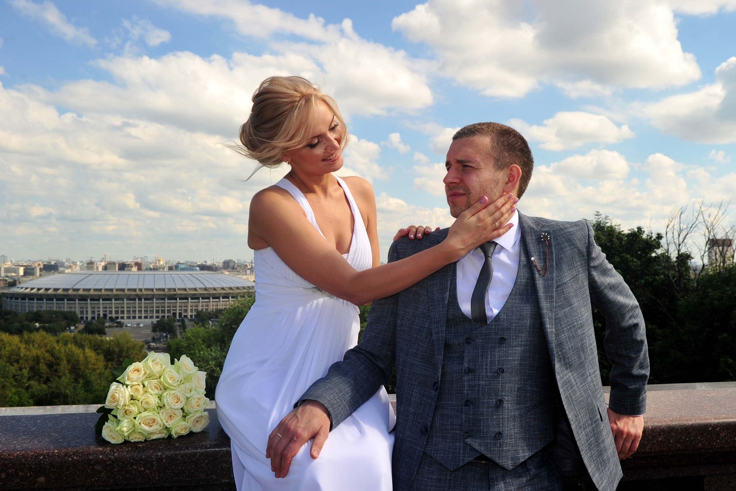 Свадьба на Воробьёвых Горах рядом с МГУ, невеста сидит на мраморном парапете смотровой площадки и нежно гладит рукой щеку жениха, жених стоит, прислонившись к ограждению смотровой площадки, на заднем плане виден стадион Лужники 