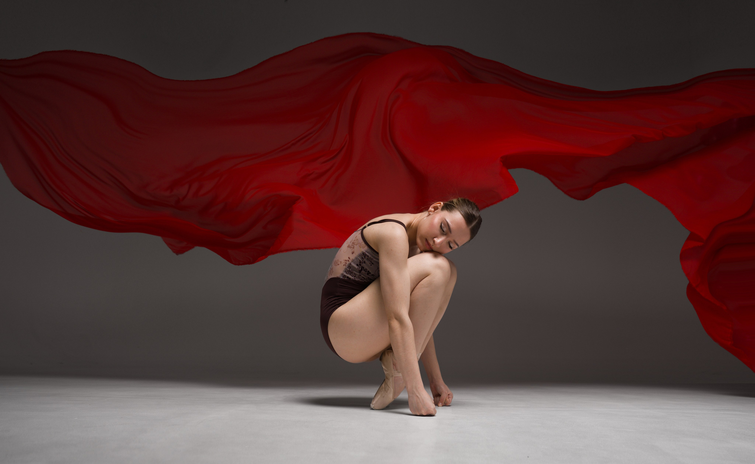 Балерина в лаконичной закрытой позе на фоне масштабной летящей красной ткани. 