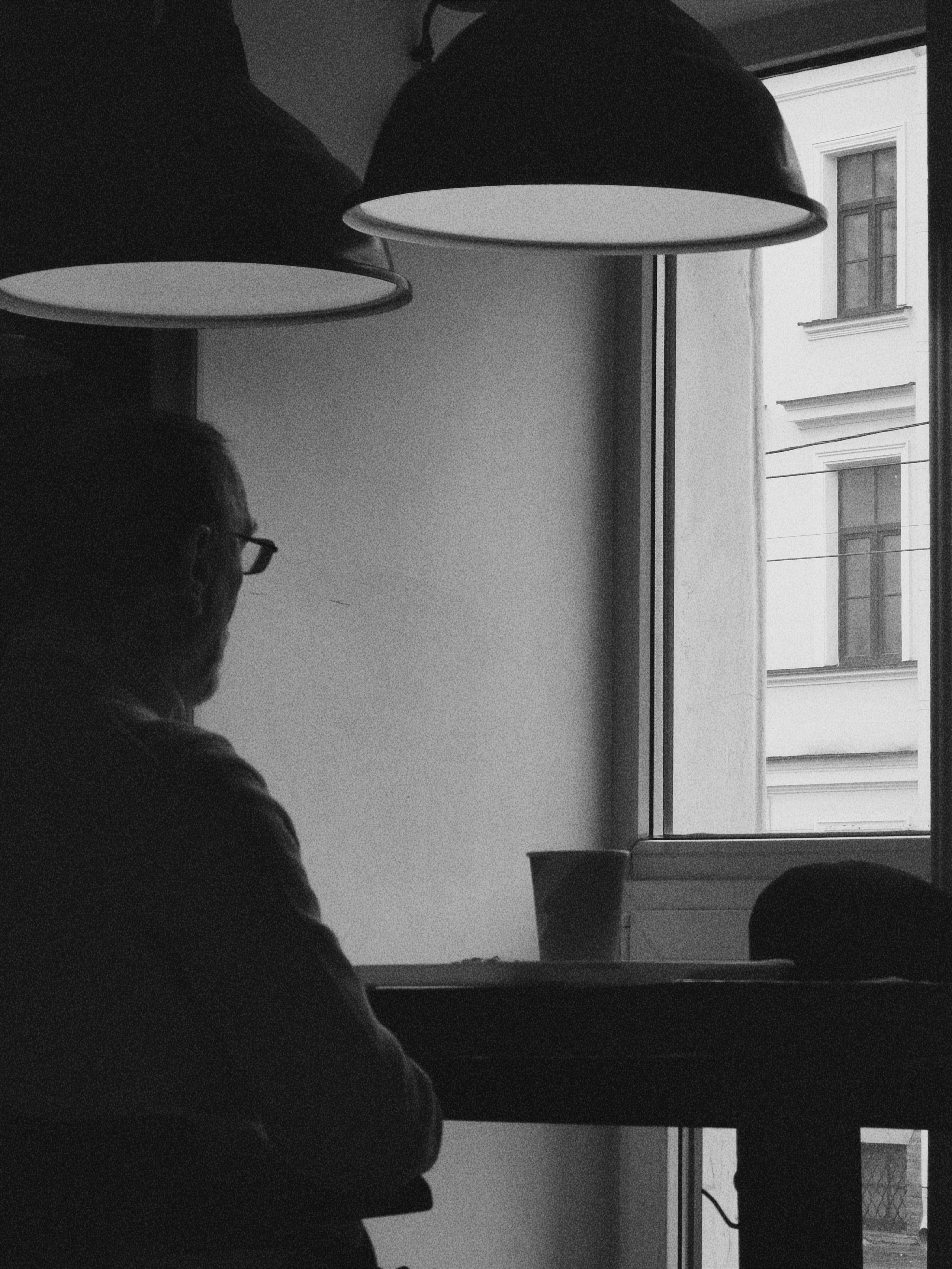 Чёрно-белая фотография: человек в очках сидит у окна в кафе, на столе стоит чашка, над ним висят подвесные лампы.
