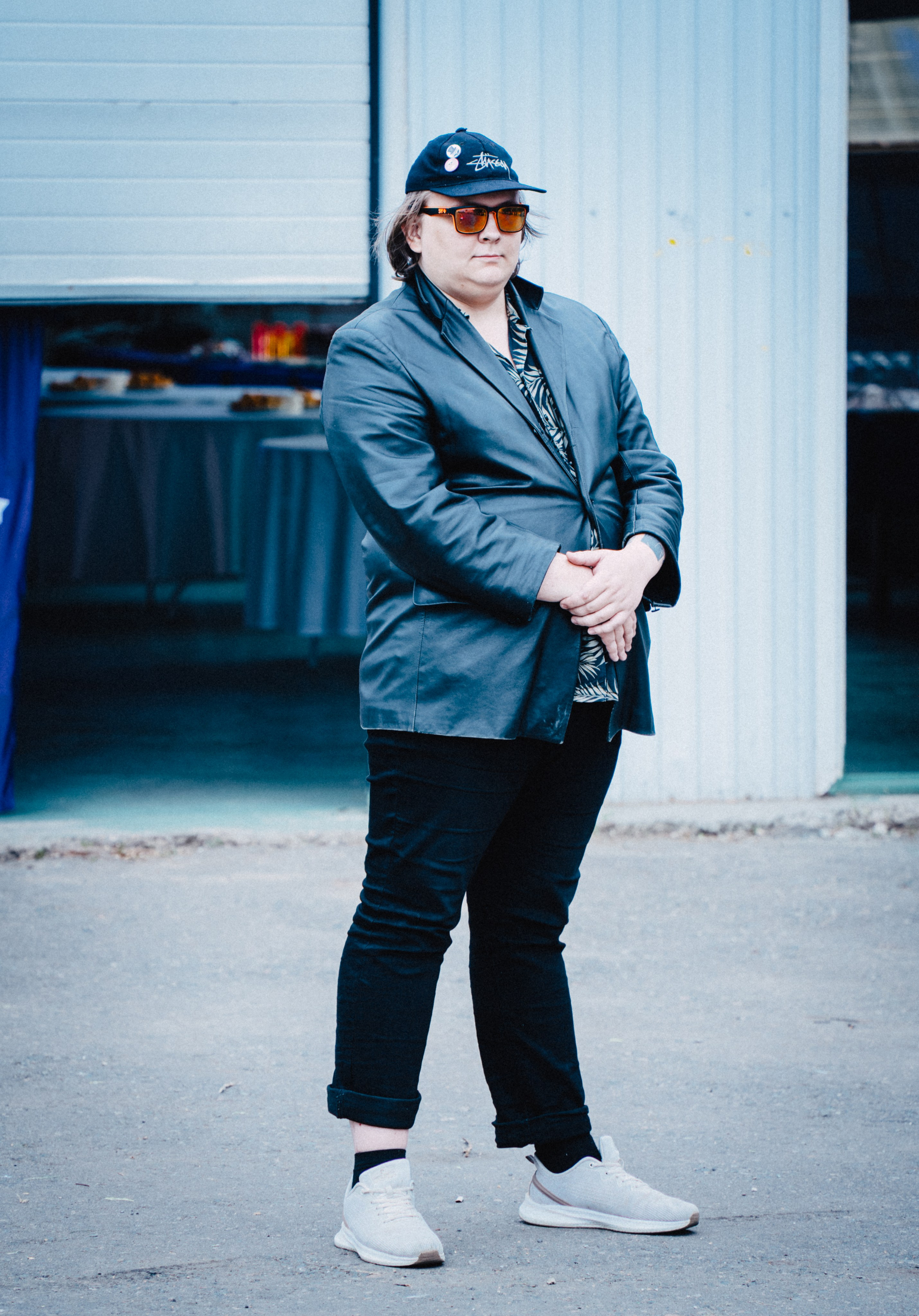 Мужчина в черной куртке, кепке и очках стоит на улице возле здания