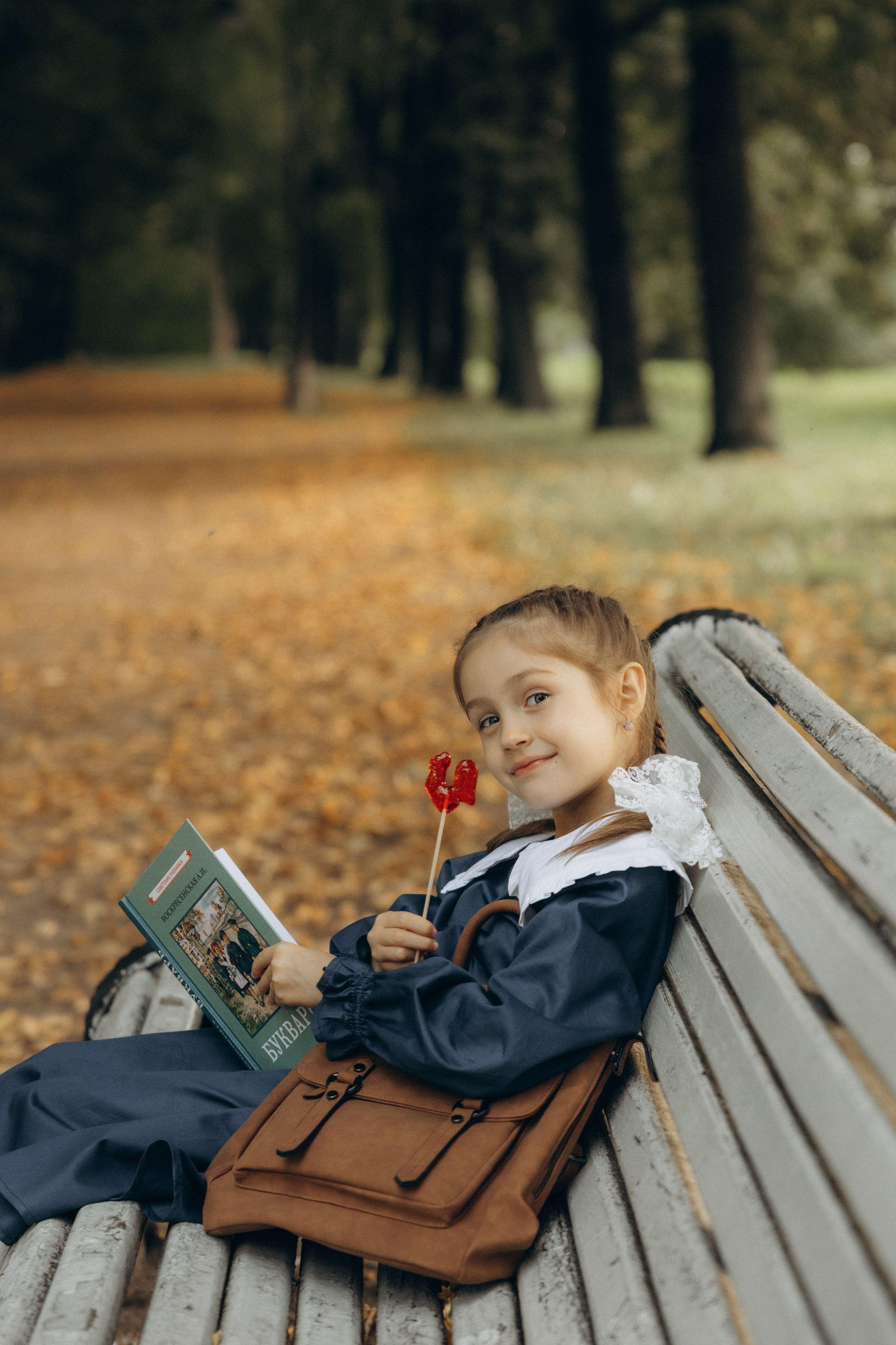 Первоклашка сидит на скамейке кушает леденец  и читает букварь