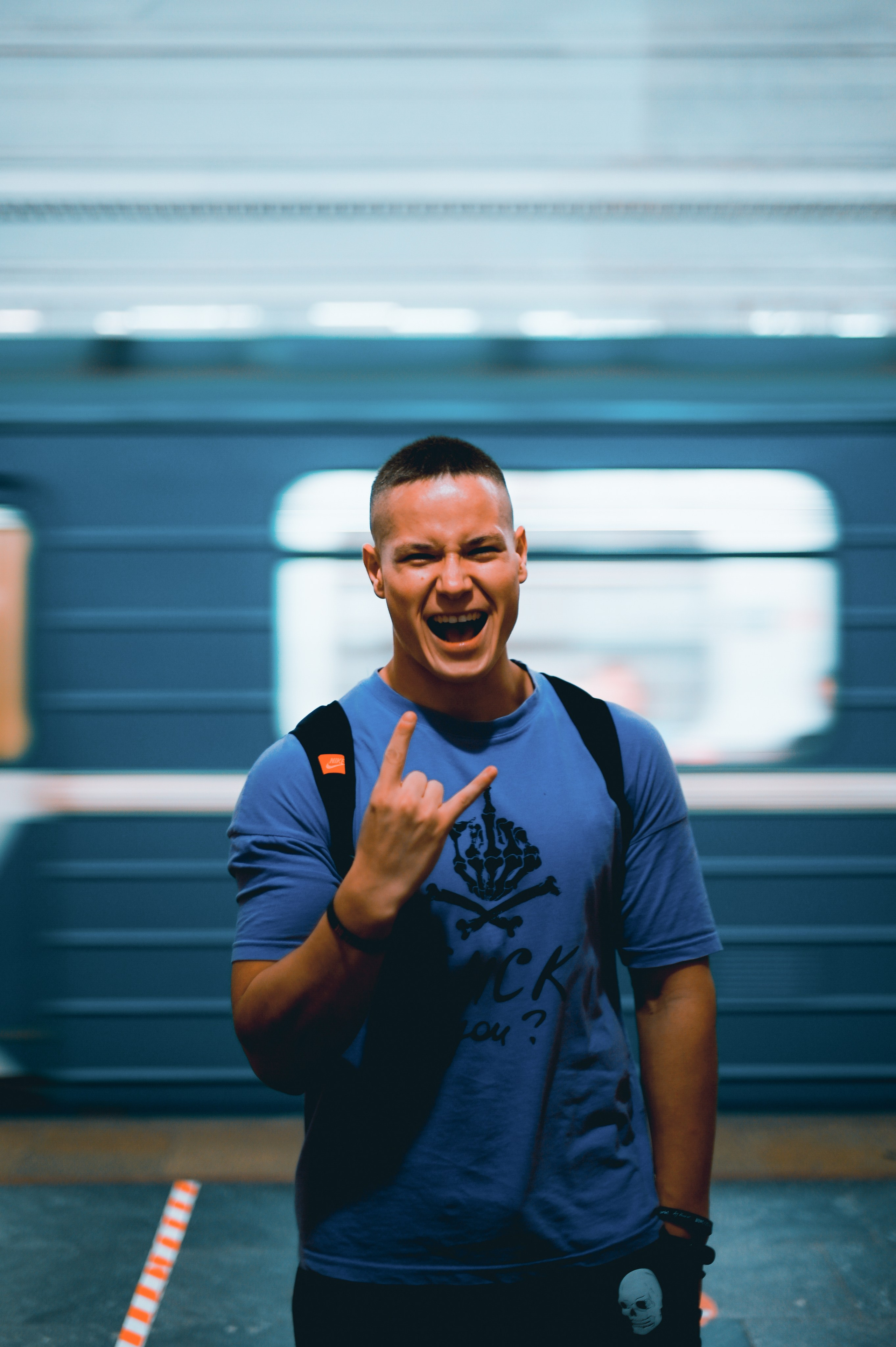 Молодой человек в синей футболке. Фон представляет собой размытое движение поезда, что создает ощущение динамики и энергии. Стильное и современное фото, отражающее уверенность и радость молодого человека.
